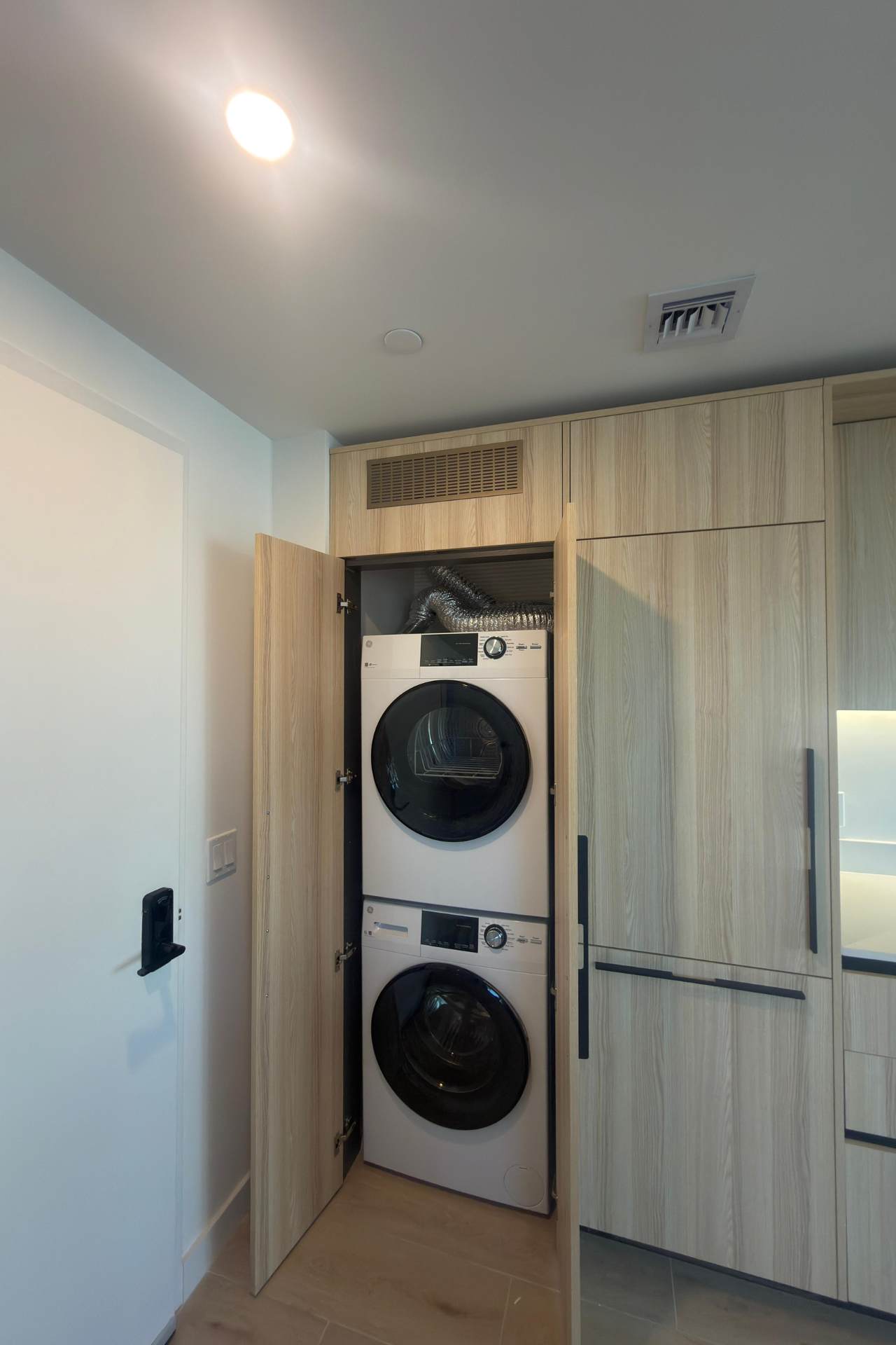 A stacked white washer and dryer are neatly tucked into a light wood-paneled closet, with the closet doors open to reveal the appliances and a ventilation hose.