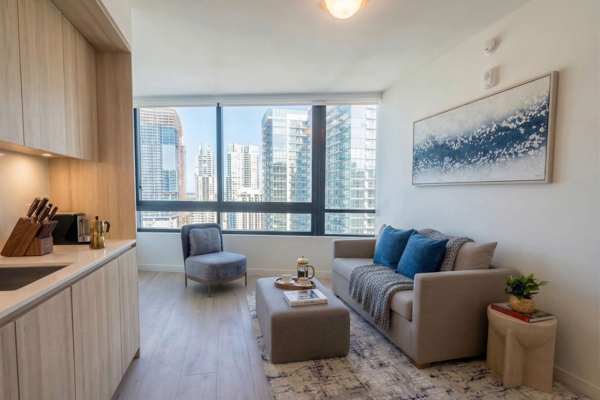 A cozy living room with a light-colored sofa, an ottoman, and a grey accent chair, all set against large windows showcasing a city skyline. A modern abstract painting hangs on the wall.