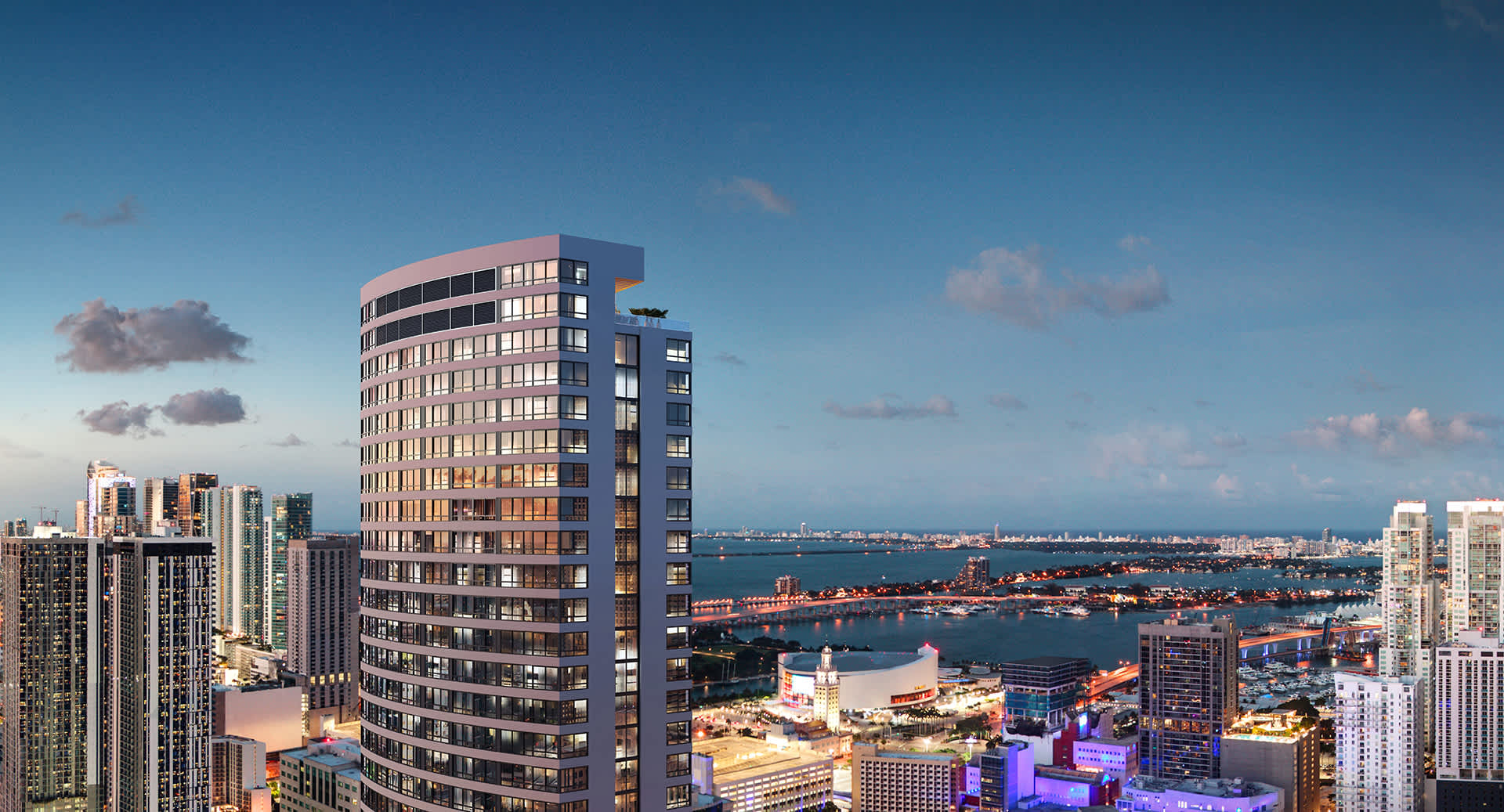 An aerial view captures a modern high-rise building with curved architecture, illuminated against a twilight sky, surrounded by a vibrant city skyline and a wide bay with distant bridges.