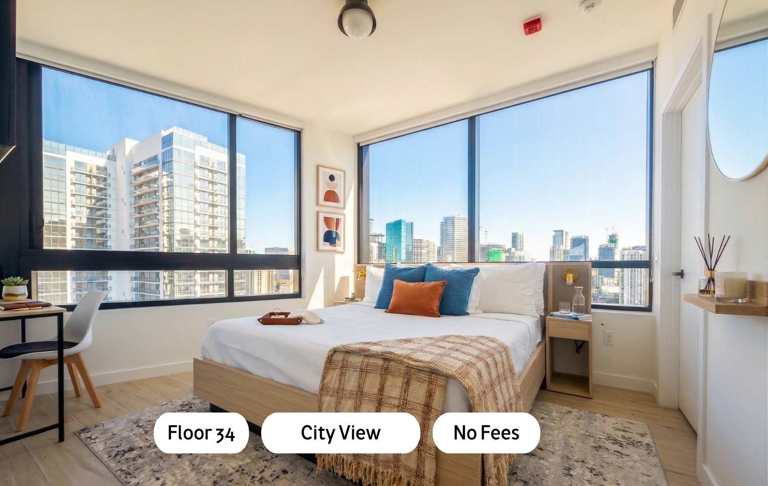 A modern bedroom with a light wood bed frame, white bedding, and colorful accent pillows, featuring large floor-to-ceiling windows offering expansive city views. A small desk and chair are visible on the left.