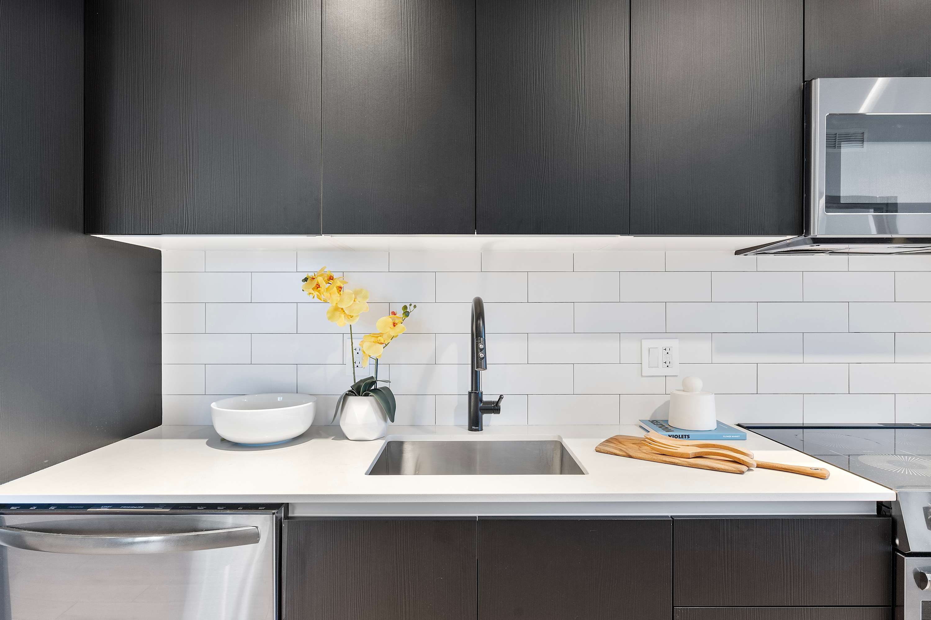 Contemporary kitchen featuring dark wood flat-panel cabinetry, white subway tile backsplash, and light quartz countertops. A stainless steel sink with a black faucet is centered, flanked by a dishwasher and microwave.