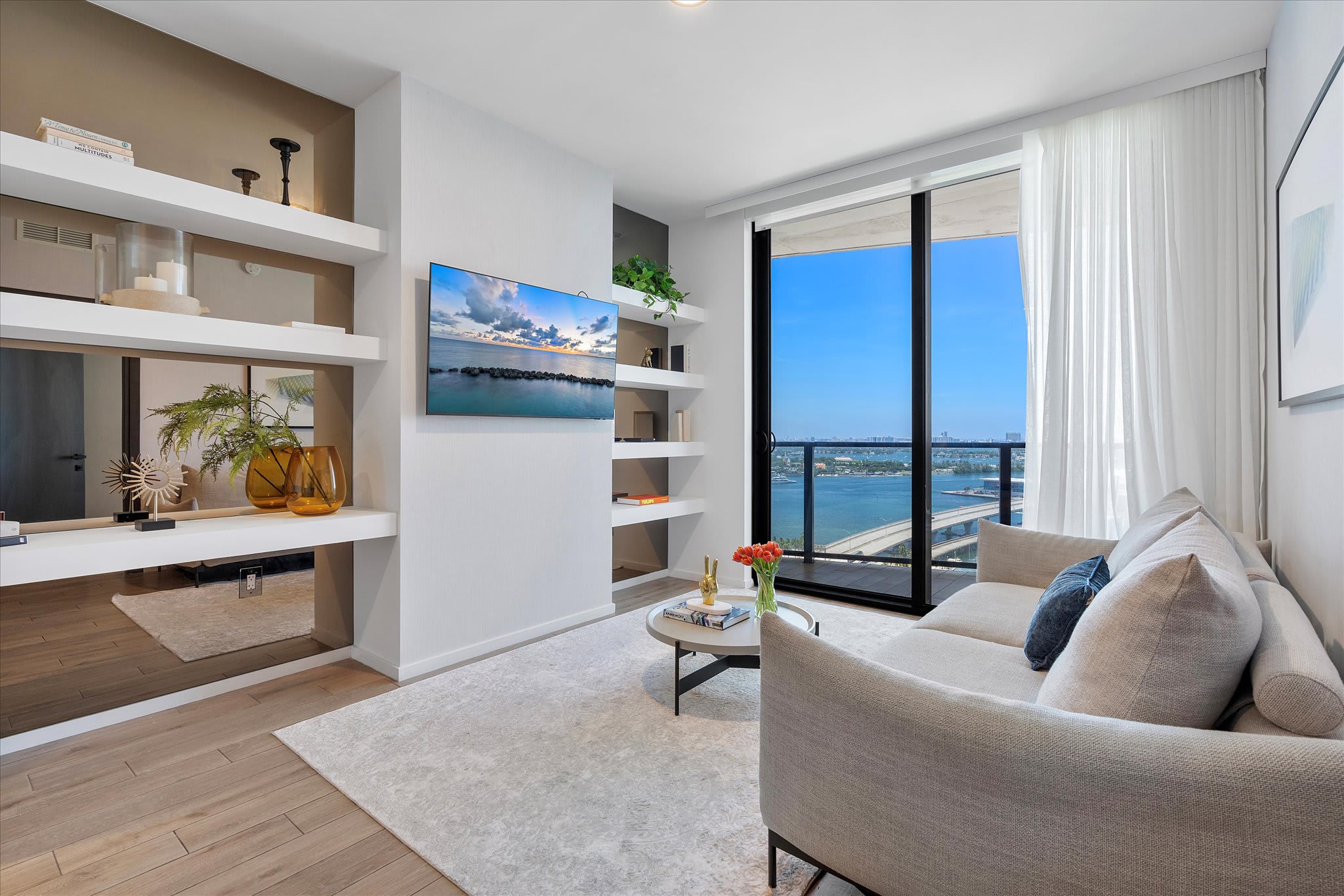 Contemporary living room with a light grey sofa, wall-mounted TV, and built-in white shelving. Floor-to-ceiling windows reveal a vibrant city and water view, enhancing the bright and airy feel.