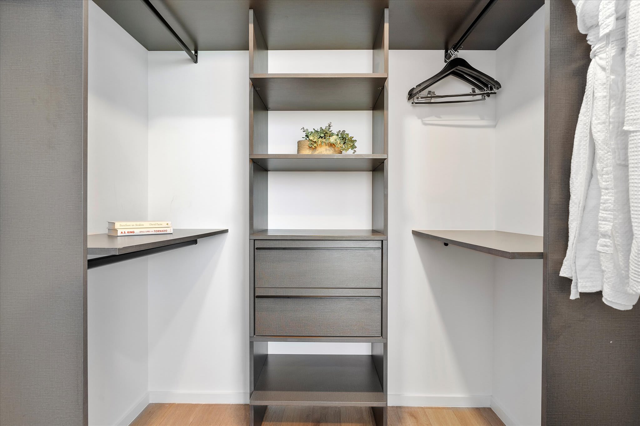 Organized walk-in closet with dark wood shelving, drawers, and hanging rods. A white bathrobe hangs on a hook, and a small plant decorates a shelf.
