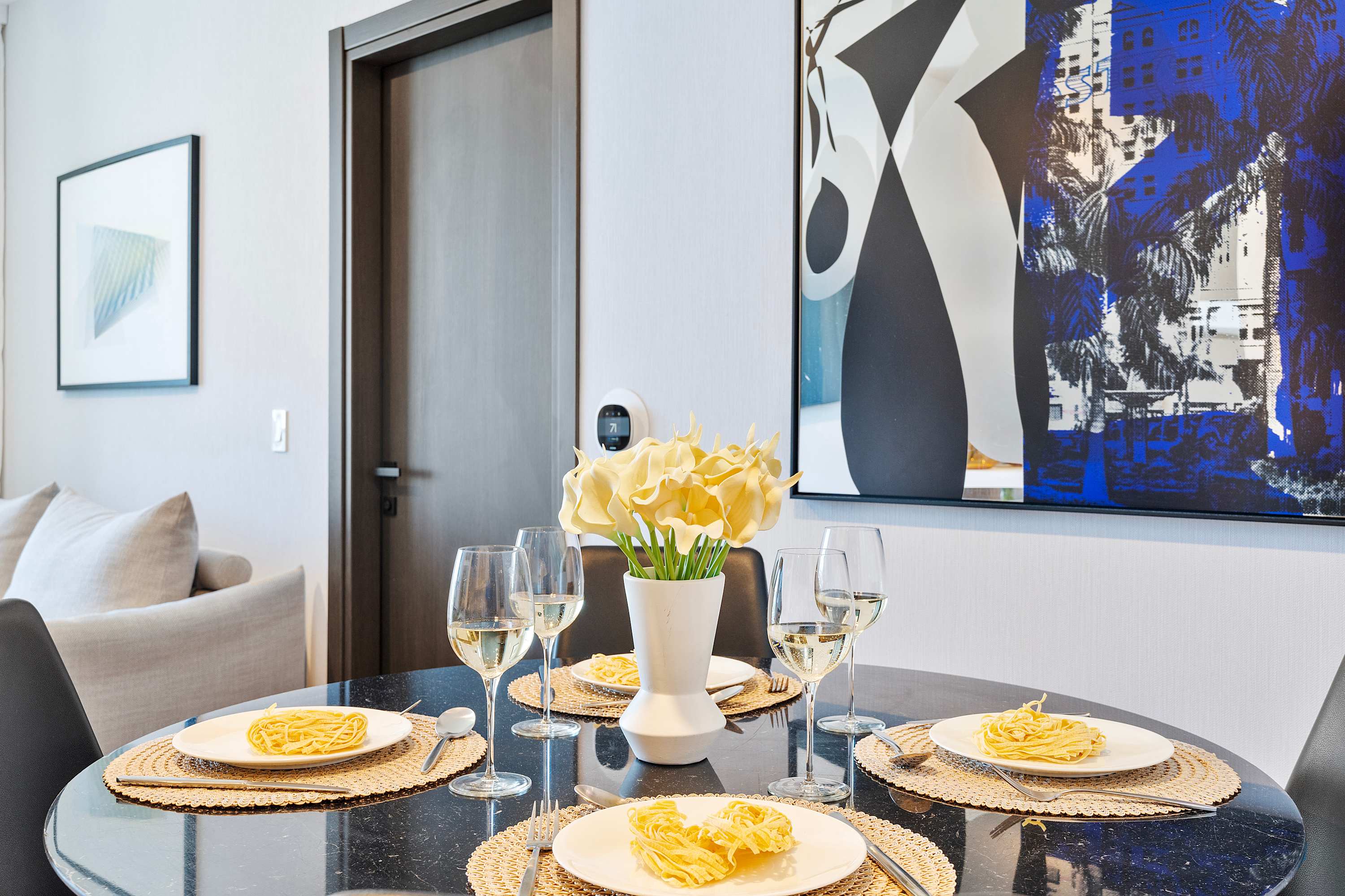 Elegant dining area with a round black marble table set for four, featuring woven placemats and white wine glasses. A vibrant abstract painting with blue and black tones adorns the wall, complementing the modern decor.