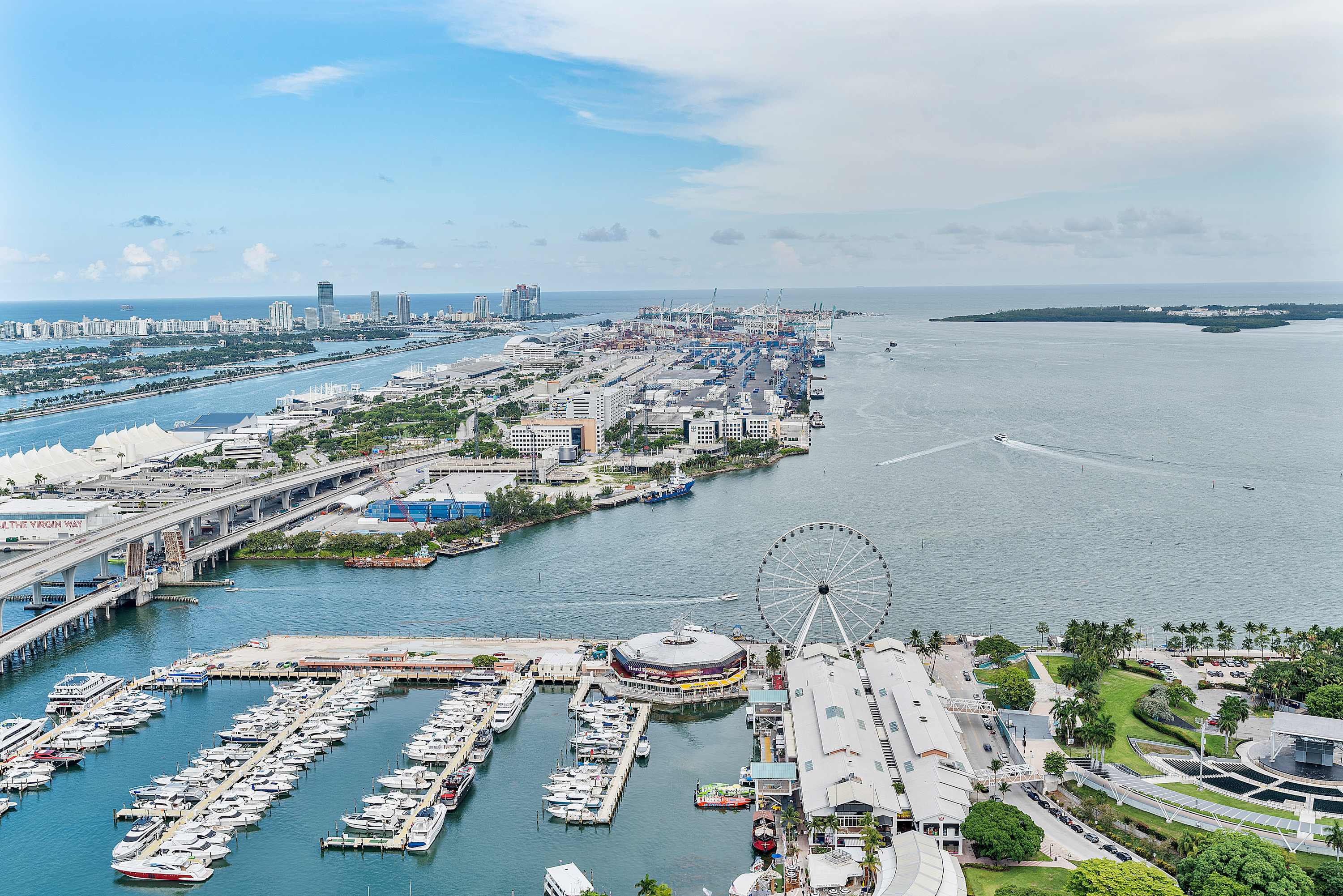 Expansive aerial view of a bustling port city with numerous cargo ships, a large Ferris wheel, and a marina filled with yachts. Bridges connect landmasses, and distant cityscapes line the horizon under a partly cloudy sky.
