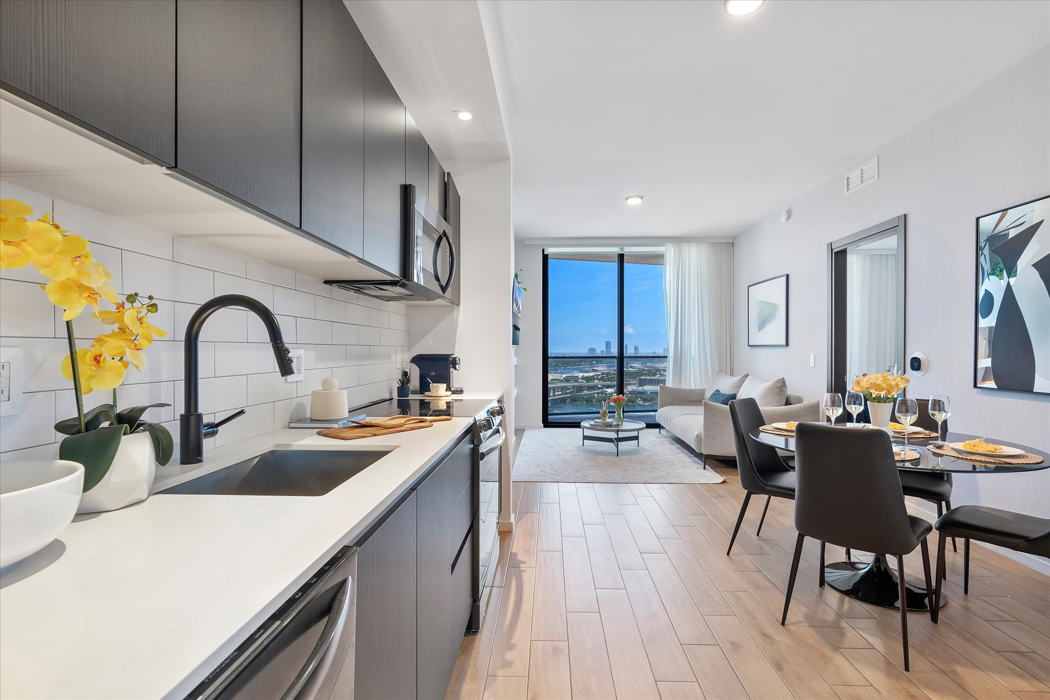 Open-concept kitchen featuring dark flat-panel cabinetry, white quartz countertops, and a subway tile backsplash, flowing into a dining area with a round black table and chairs, and a living room with a balcony view of the city and water.