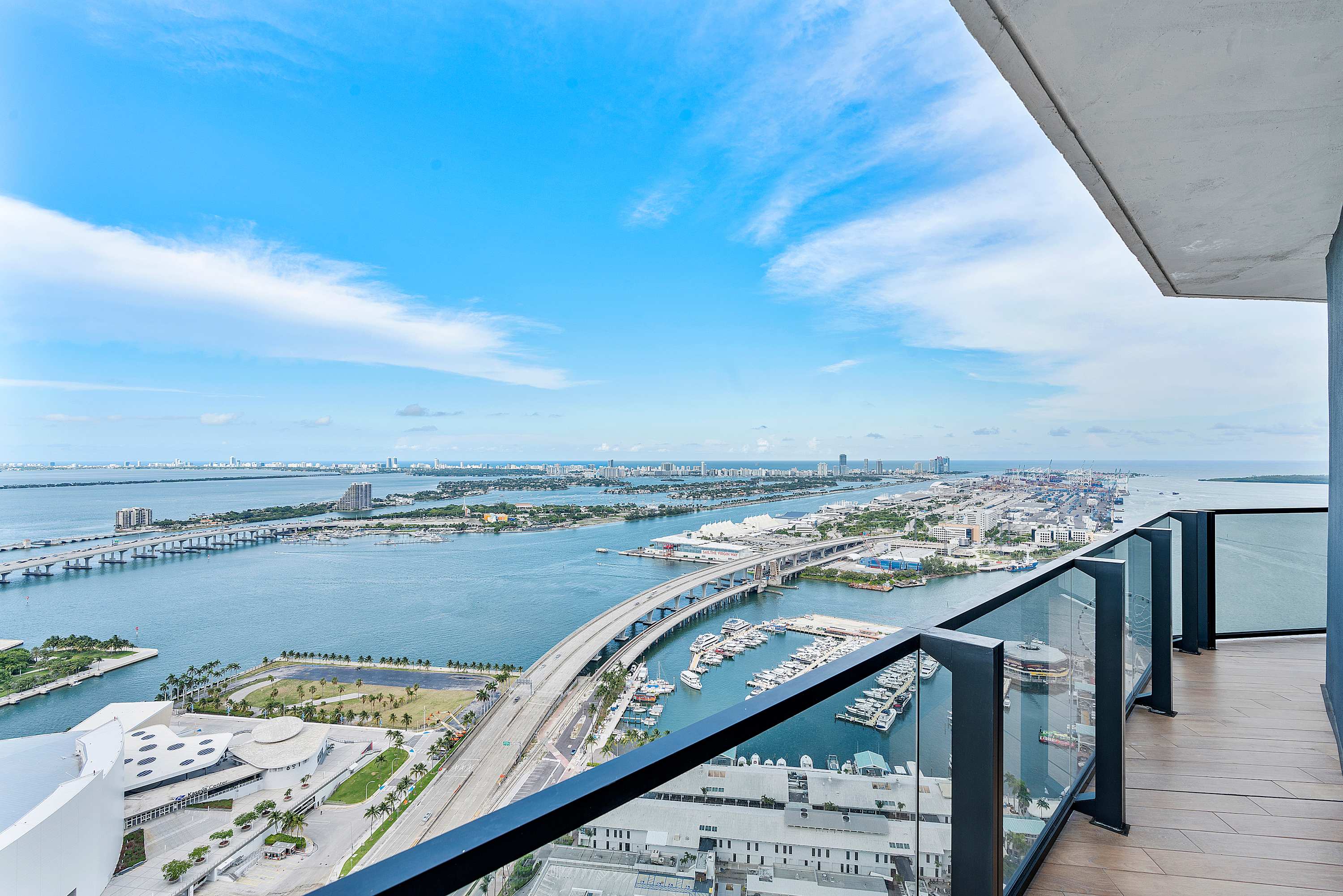 Expansive private balcony with glass railings offering a breathtaking panoramic view of the ocean, bay, and city skyline, including multiple bridges and a marina under a clear blue sky.