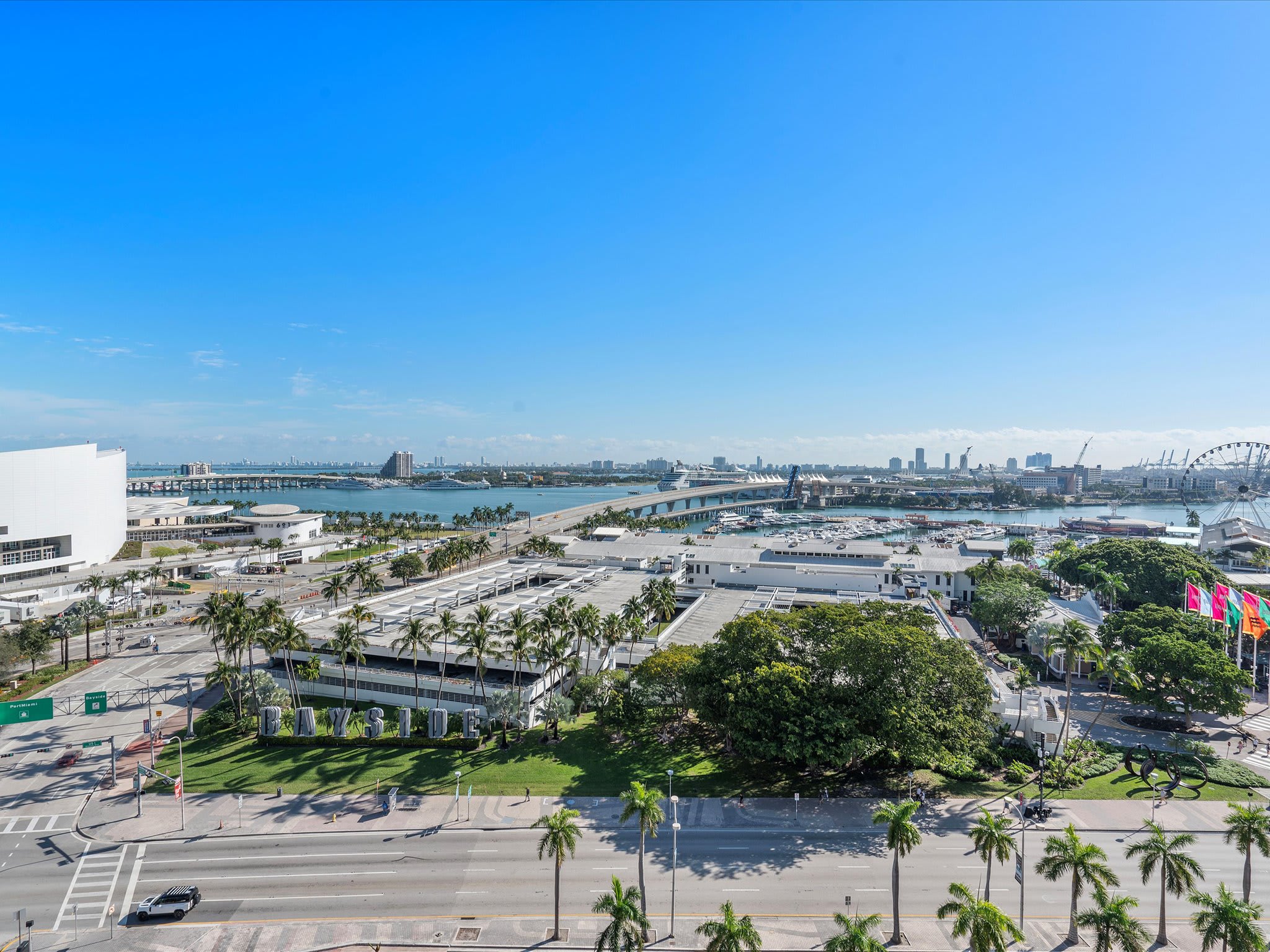A panoramic aerial view of a vibrant city waterfront, featuring a wide road lined with palm trees, a large parking structure, and a bustling bay with bridges and cruise ships, all under a clear blue sky.