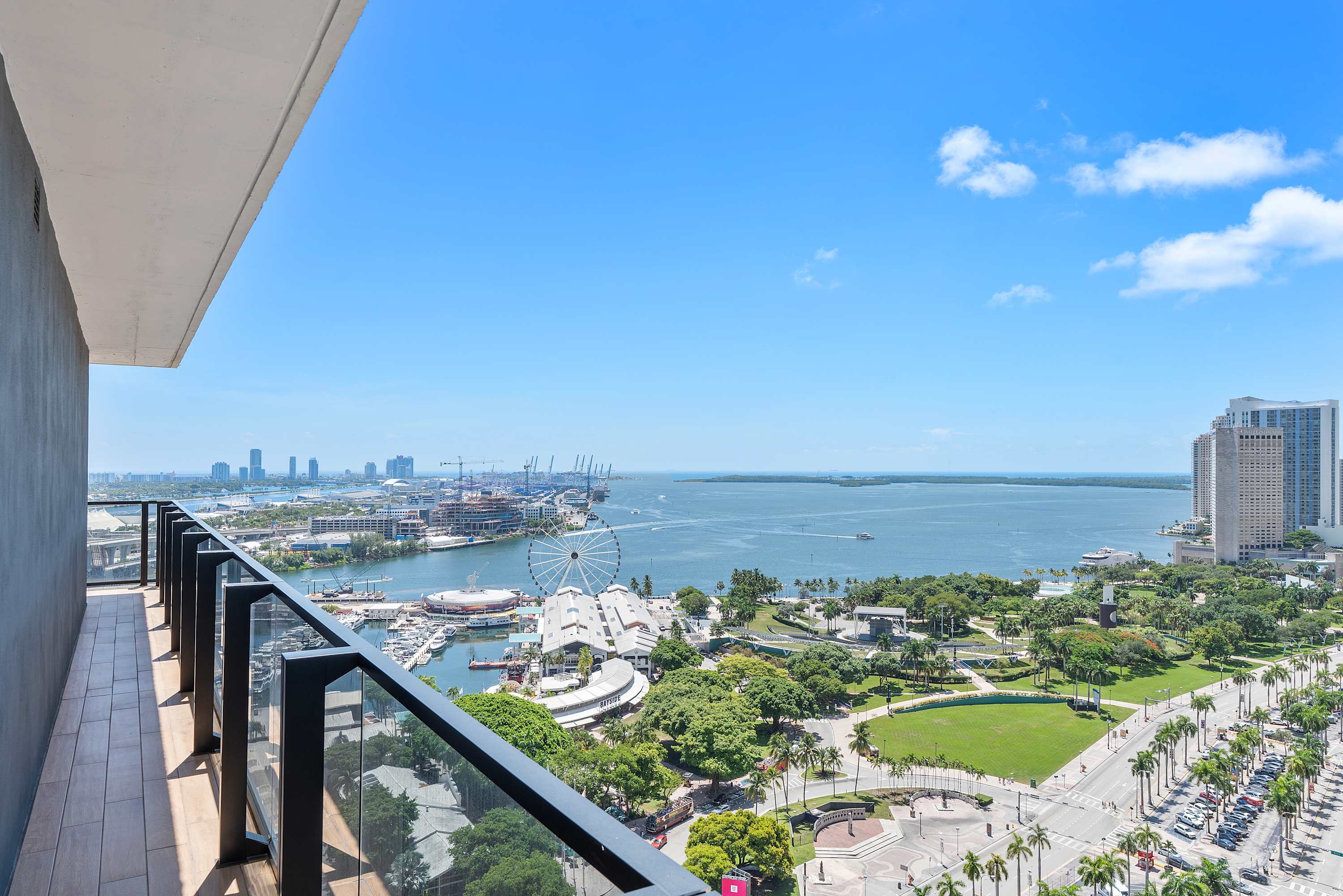 High-angle view from a balcony with glass railings, showcasing a vibrant city skyline, a large bay with boats, and a prominent Ferris wheel under a clear blue sky. The foreground features a wooden-decked balcony.