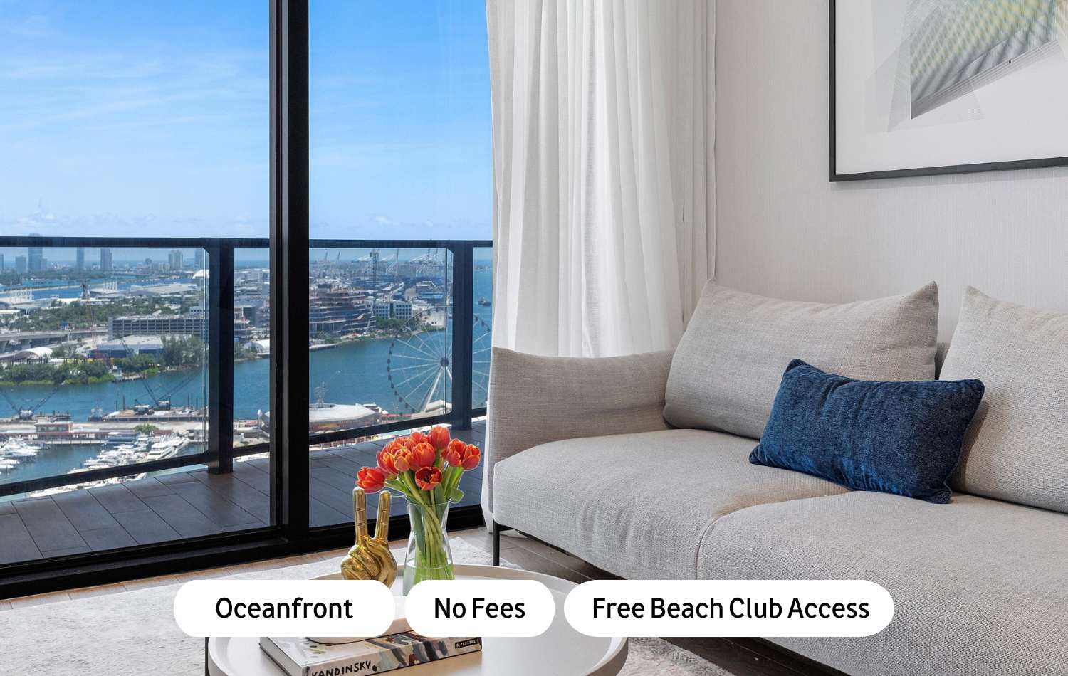 Modern living room with a light grey sofa, blue accent pillow, and a white coffee table with orange tulips. Floor-to-ceiling windows offer panoramic ocean and city views, including a Ferris wheel and marina.