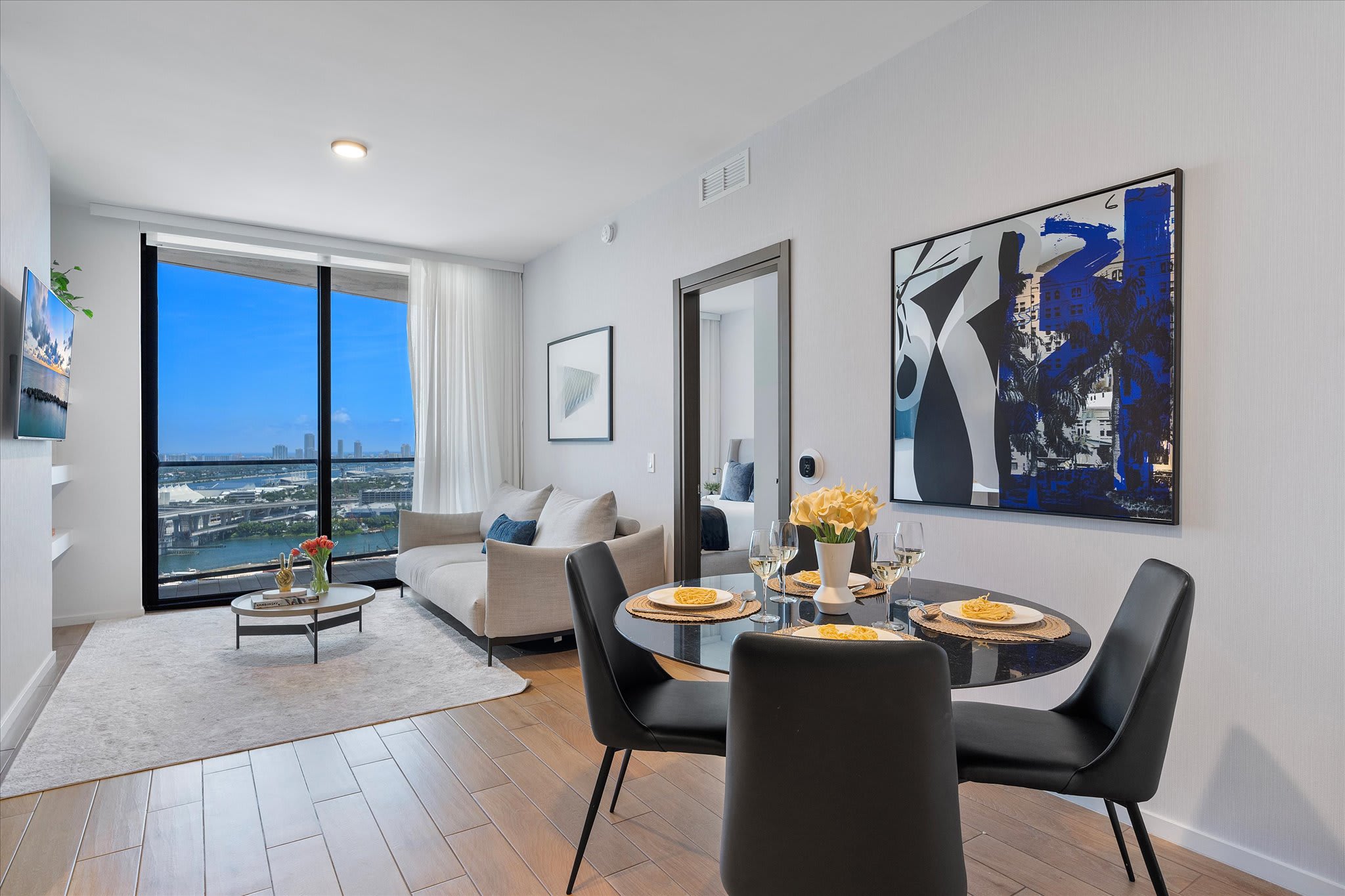 Open-concept living and dining area with a light grey sofa, a round black dining table with four chairs, and a large abstract painting. Floor-to-ceiling windows provide expansive city and water views.