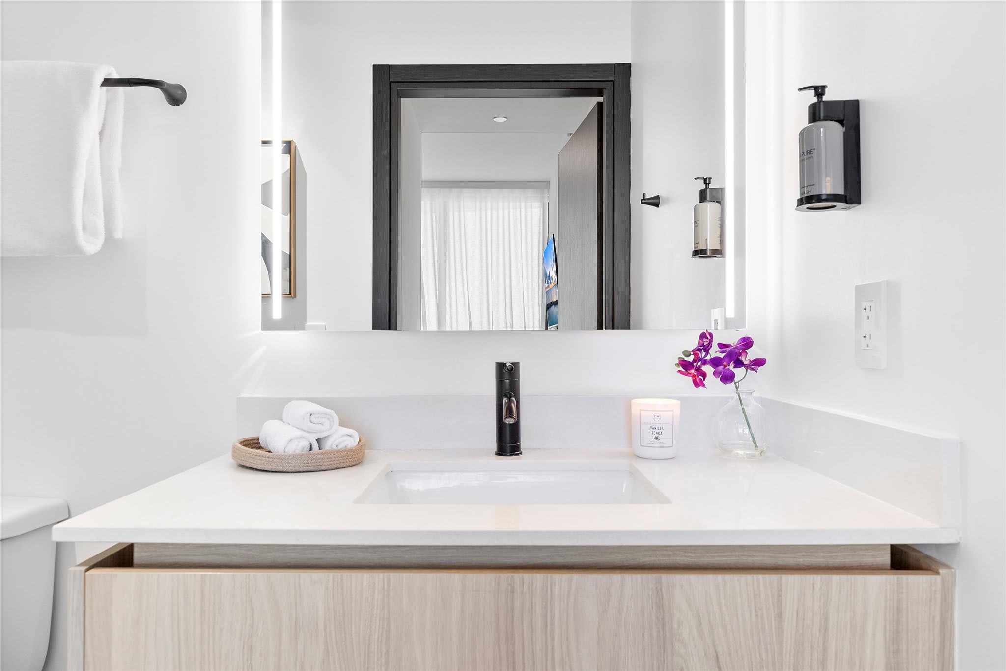 Contemporary bathroom vanity with a light wood cabinet, white countertop, and a rectangular sink with a black faucet. An illuminated mirror hangs above, and a small orchid decorates the counter.