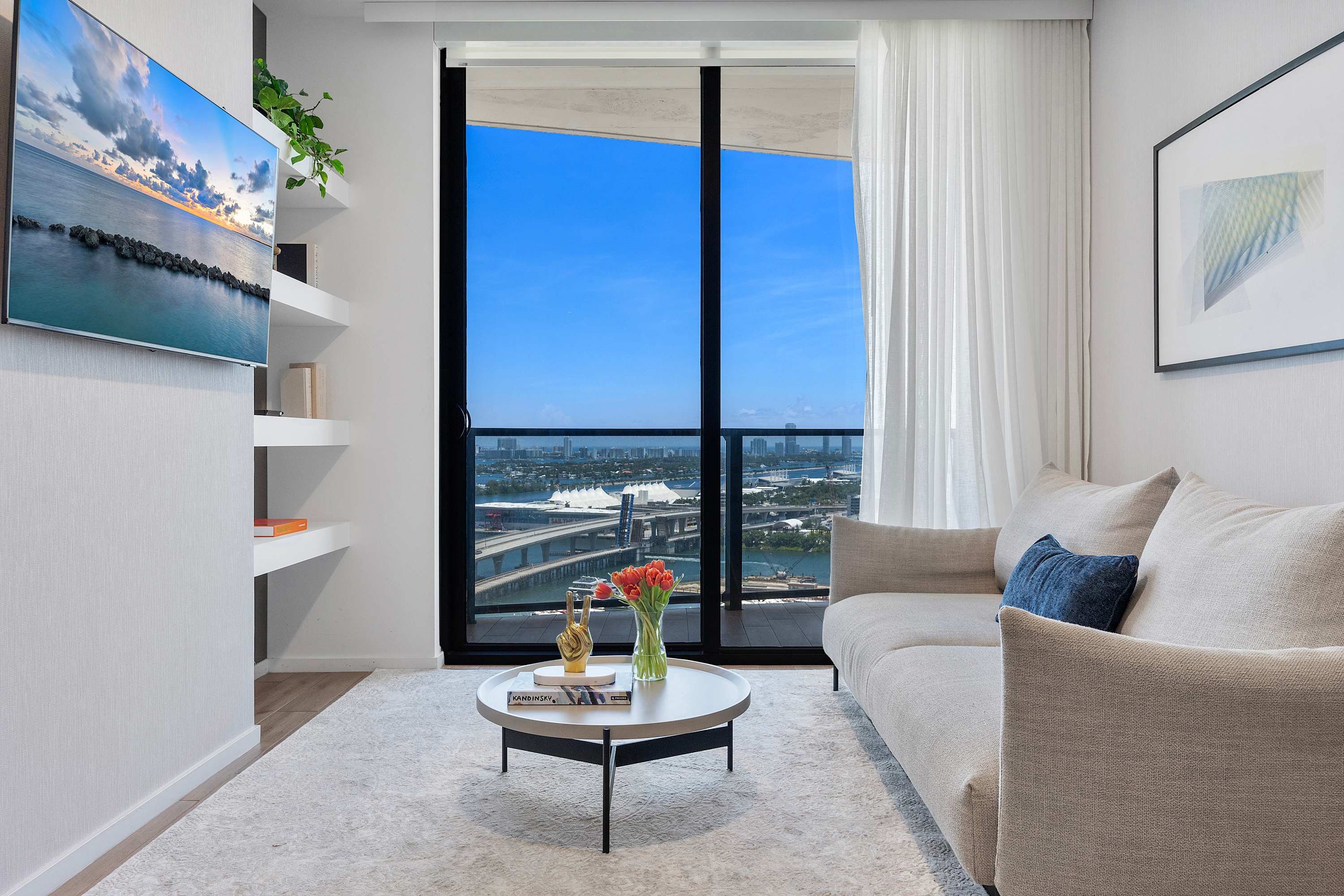 Modern living room with a light grey sofa, white coffee table, and a wall-mounted TV. Floor-to-ceiling windows provide expansive views of the city and water, with sheer white curtains adding softness.