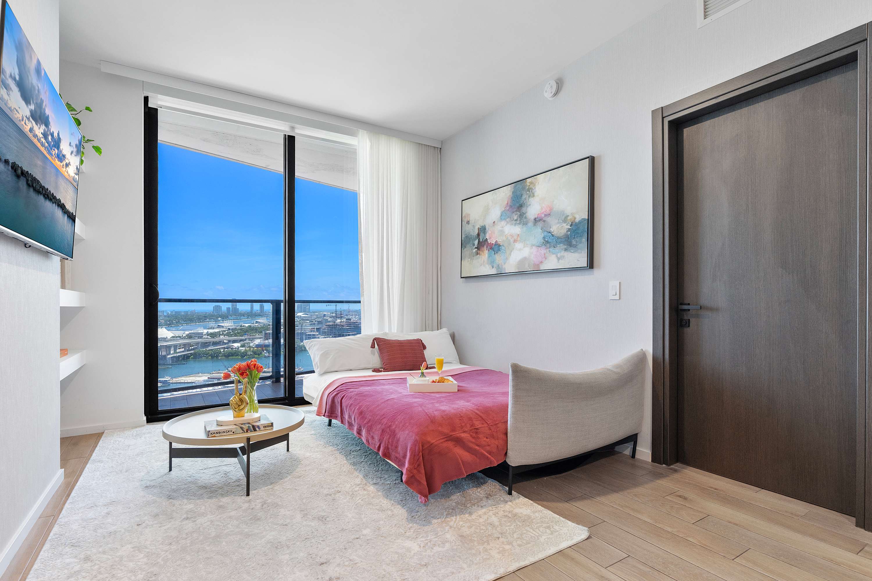 Bright bedroom, potentially a convertible space, with a bed featuring a pink throw and a tray with breakfast. Floor-to-ceiling windows offer a stunning view of the city and water, with a modern abstract painting on the wall.