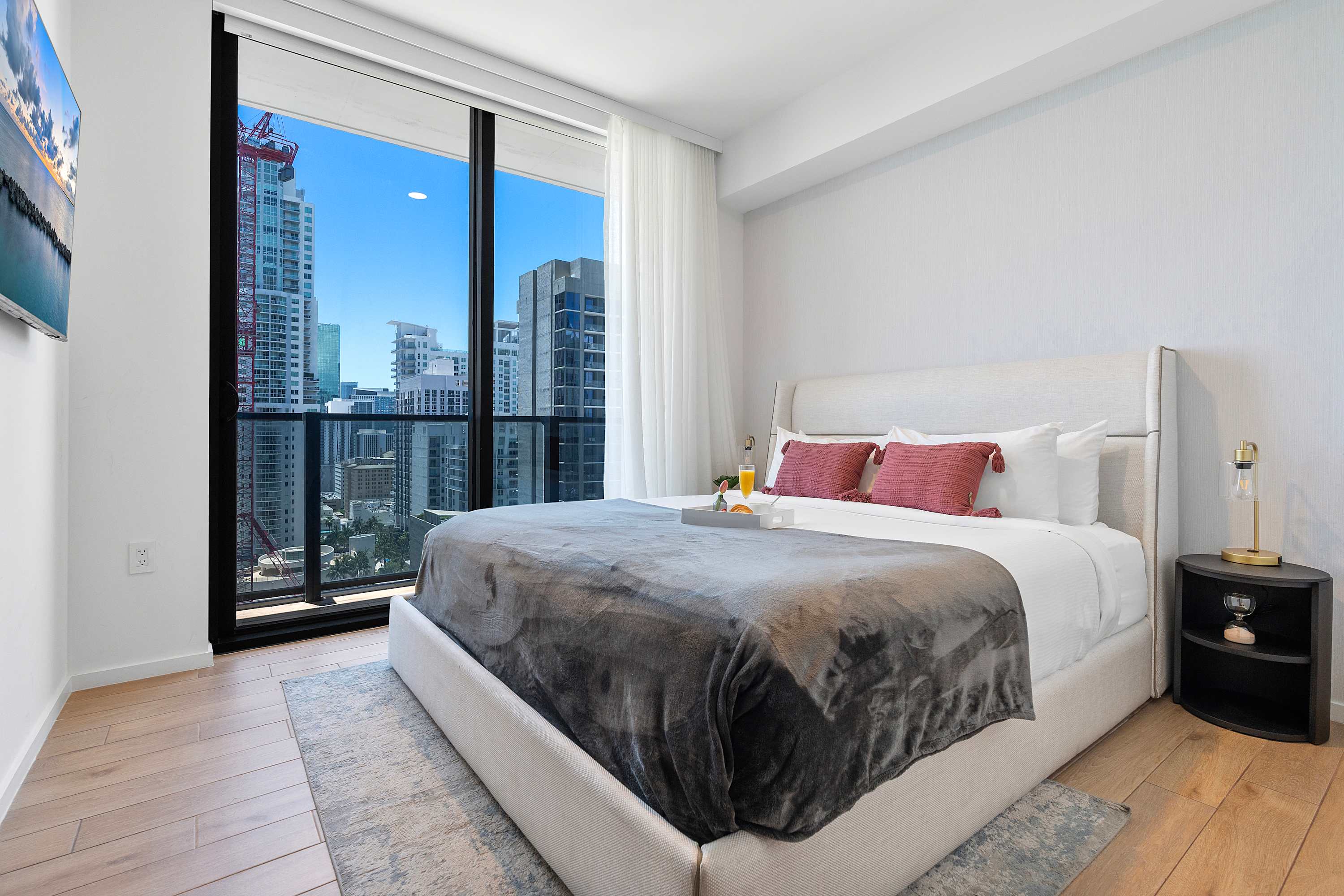 Bright bedroom with a king-sized bed featuring a light upholstered headboard and grey bedding. Floor-to-ceiling windows offer a stunning city skyline view with modern high-rise buildings.