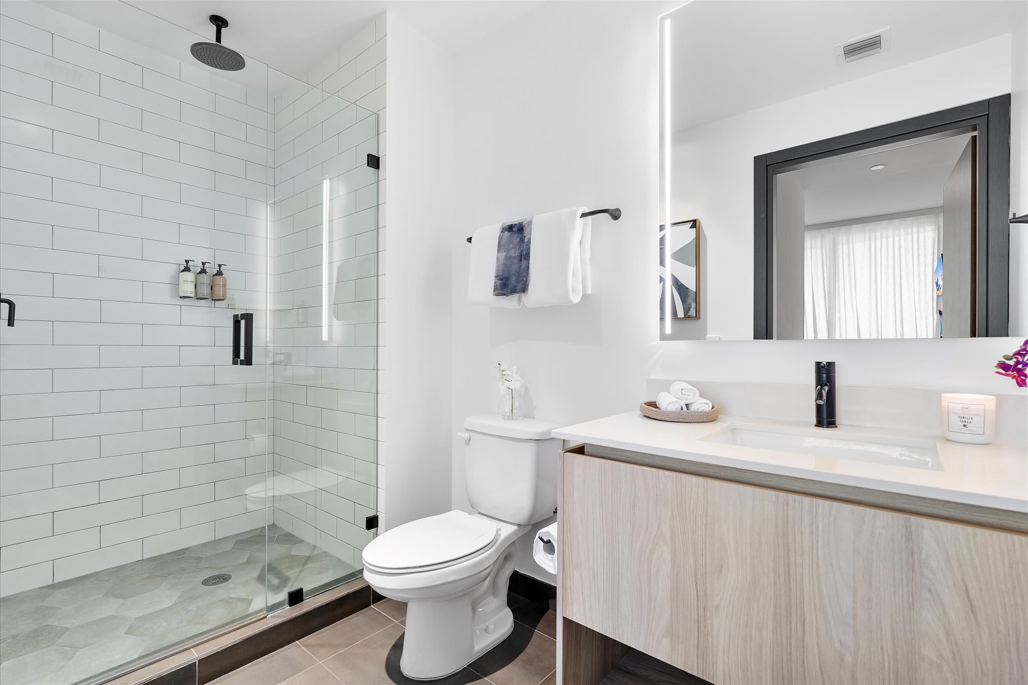 Modern bathroom with a white subway tile shower, glass enclosure, and dark hexagonal floor tiles. A white toilet and a light wood vanity with a white countertop and illuminated mirror are also visible.