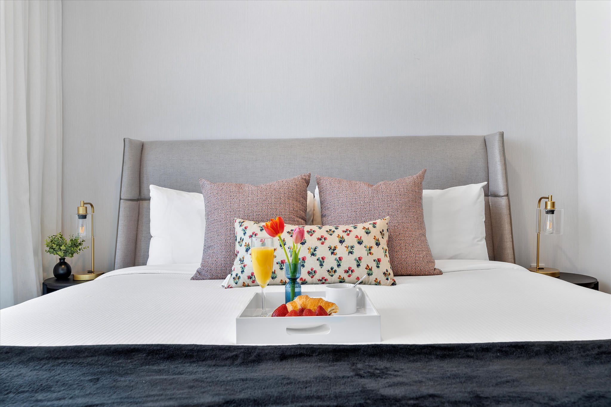 Cozy bedroom featuring a large gray upholstered headboard, white bedding with decorative pillows, and two modern bedside lamps. A breakfast tray with juice and a croissant is placed on the bed.