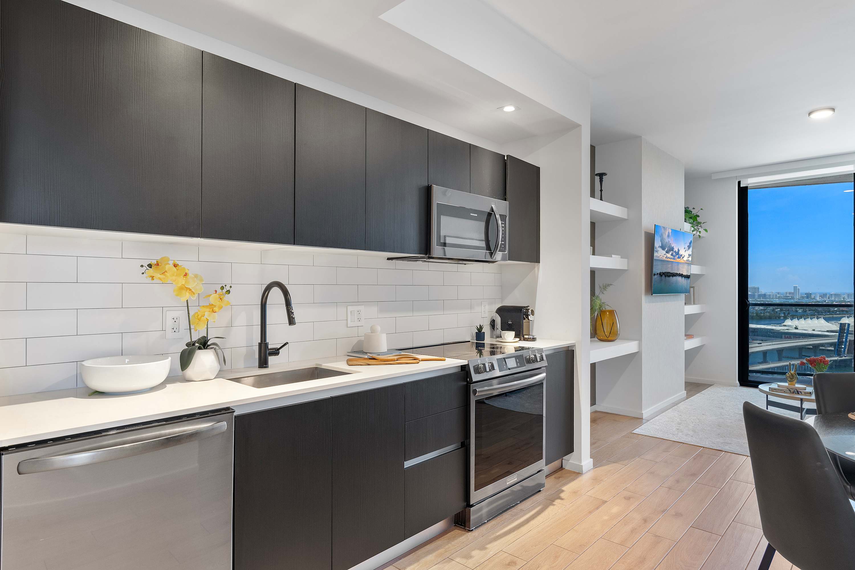 Modern kitchen with dark wood flat-panel cabinets, white subway tile backsplash, white quartz countertops, and stainless steel appliances including a dishwasher, sink, microwave, and range. A yellow orchid adds a pop of color.