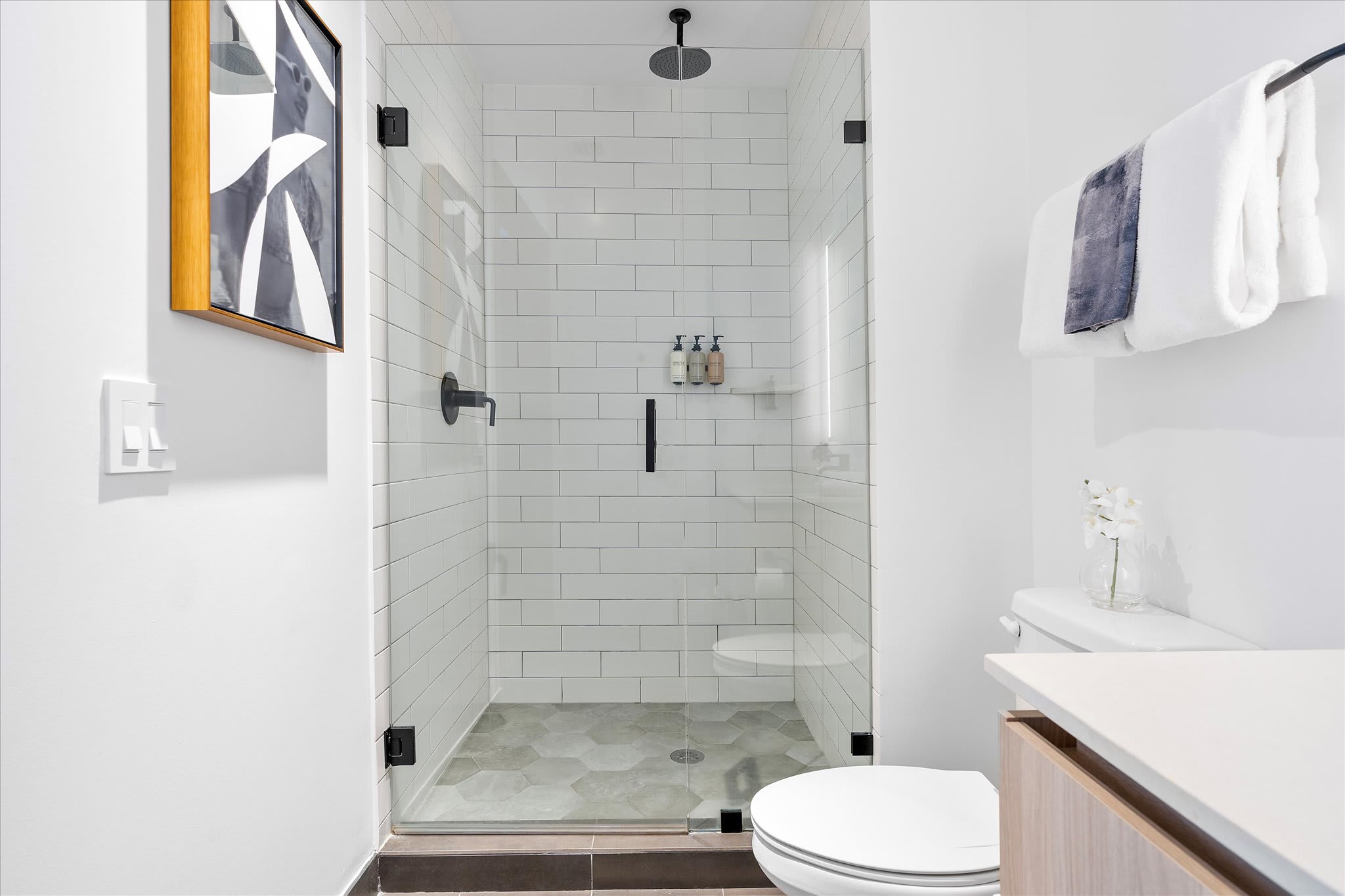 Close-up of a modern shower with white subway tiles, a glass enclosure, and a dark hexagonal tile floor. Black fixtures, including a rain shower head and a wall-mounted caddy with toiletries, are featured.