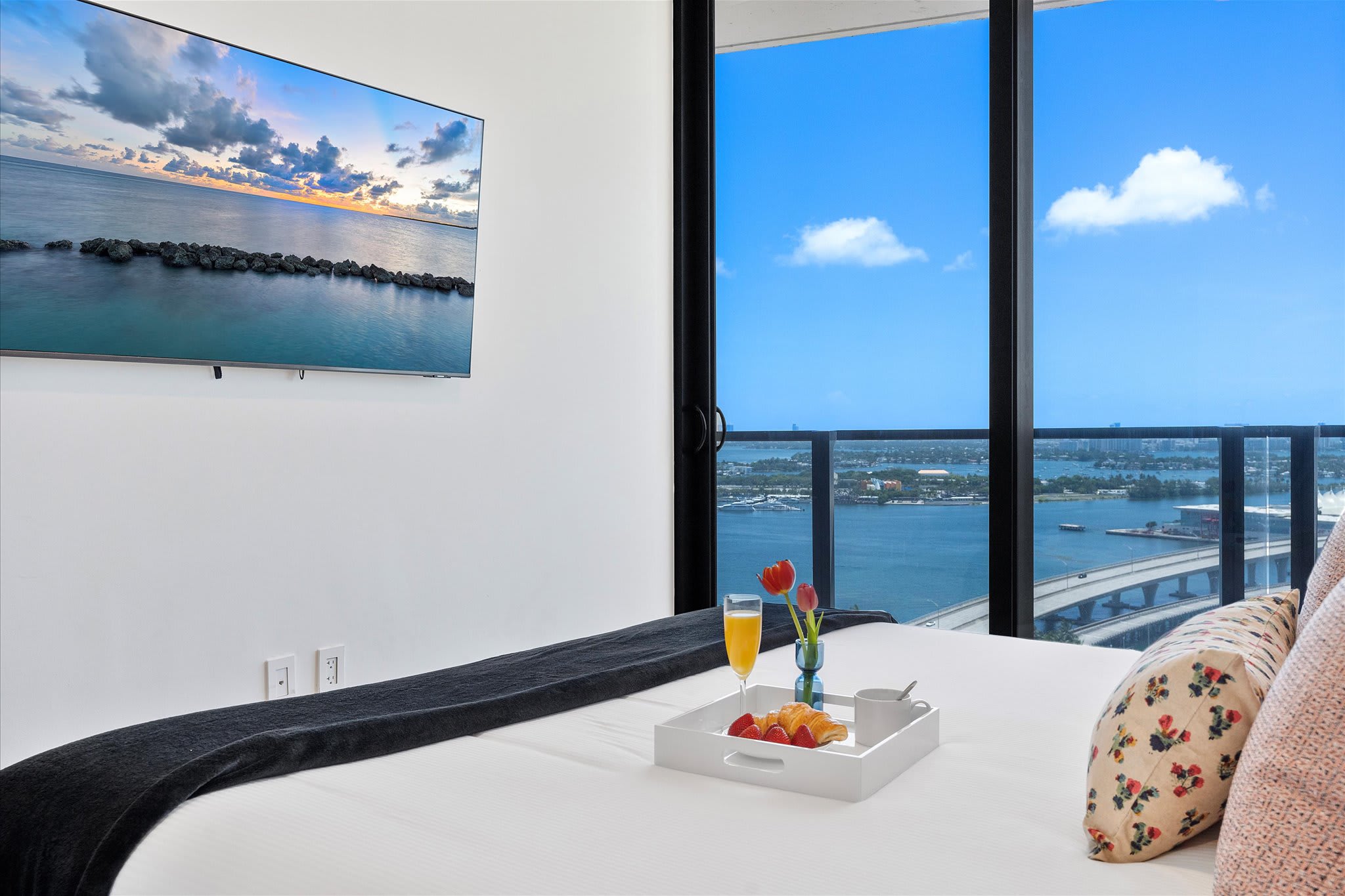 Bedroom with a large bed and a wall-mounted TV, offering expansive views of the city and water through floor-to-ceiling glass doors leading to a balcony. A breakfast tray is on the bed.