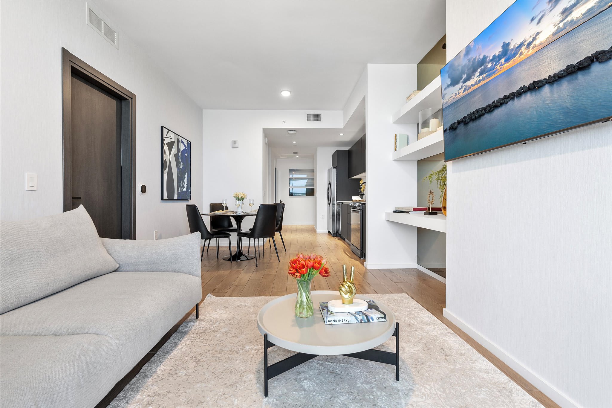 Bright living room with light hardwood floors, a light gray sofa, and a round coffee table with a vase of red tulips. A black dining table with chairs is visible in the background, and a large TV is mounted on the wall.