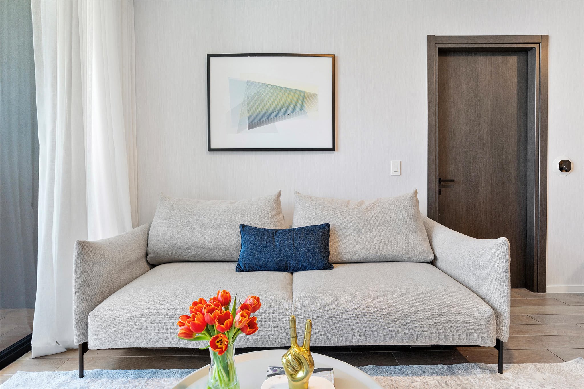 Cozy living room featuring a light grey sofa with a blue accent pillow, a white round coffee table, and a framed abstract artwork. A dark wooden door is visible in the background.