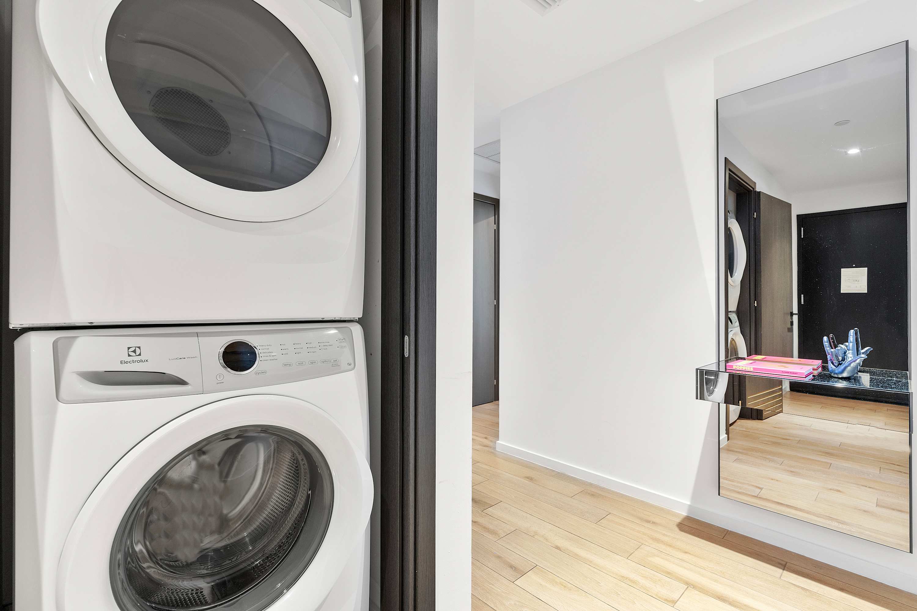Stacked white Electrolux washer and dryer unit located in a hallway nook with light hardwood floors. A full-length mirror and a small console table are visible nearby.