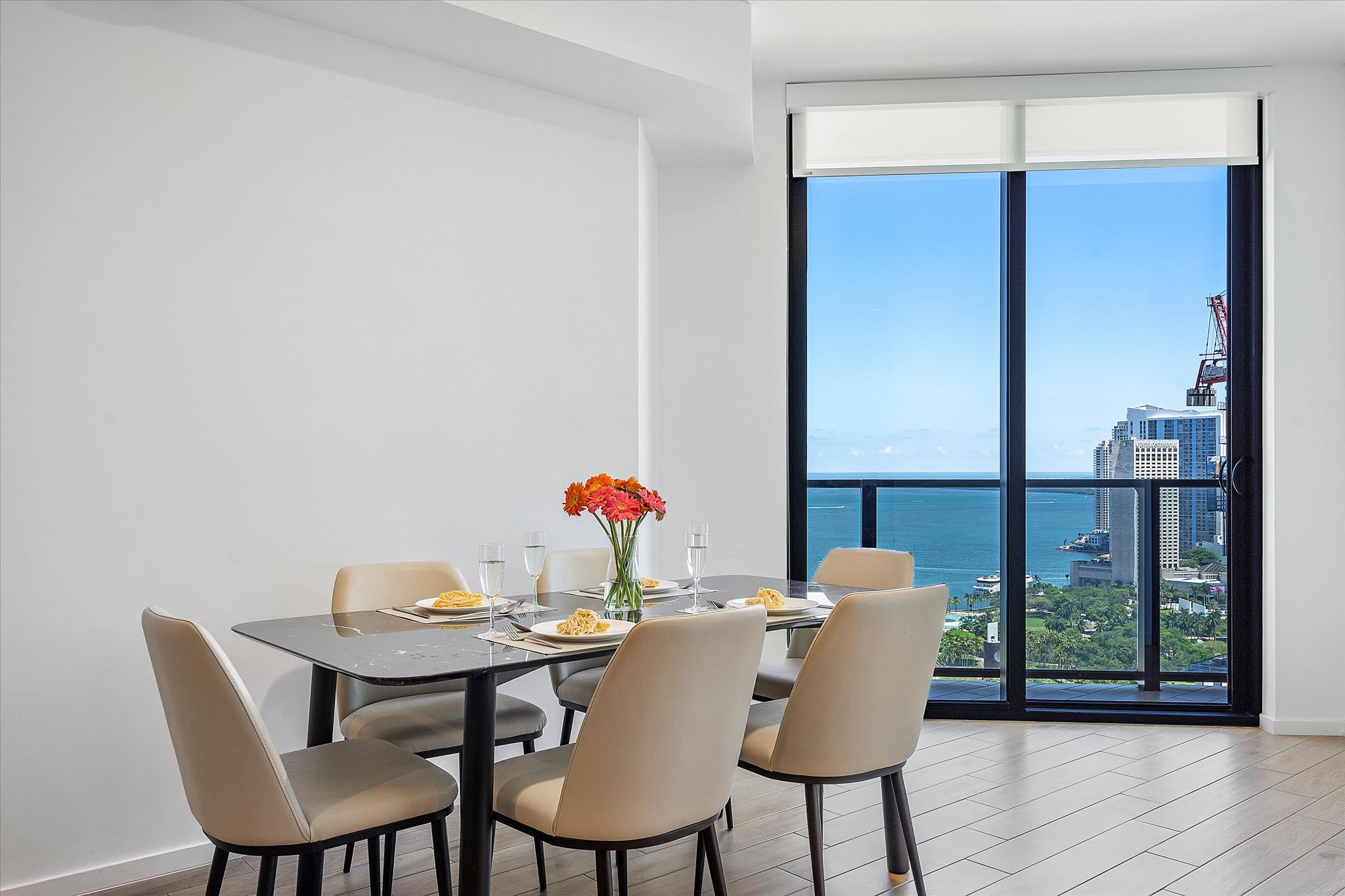 Elegant dining area featuring a dark marble-top table with four light-colored upholstered chairs, positioned next to a large floor-to-ceiling window offering panoramic city and bay views.