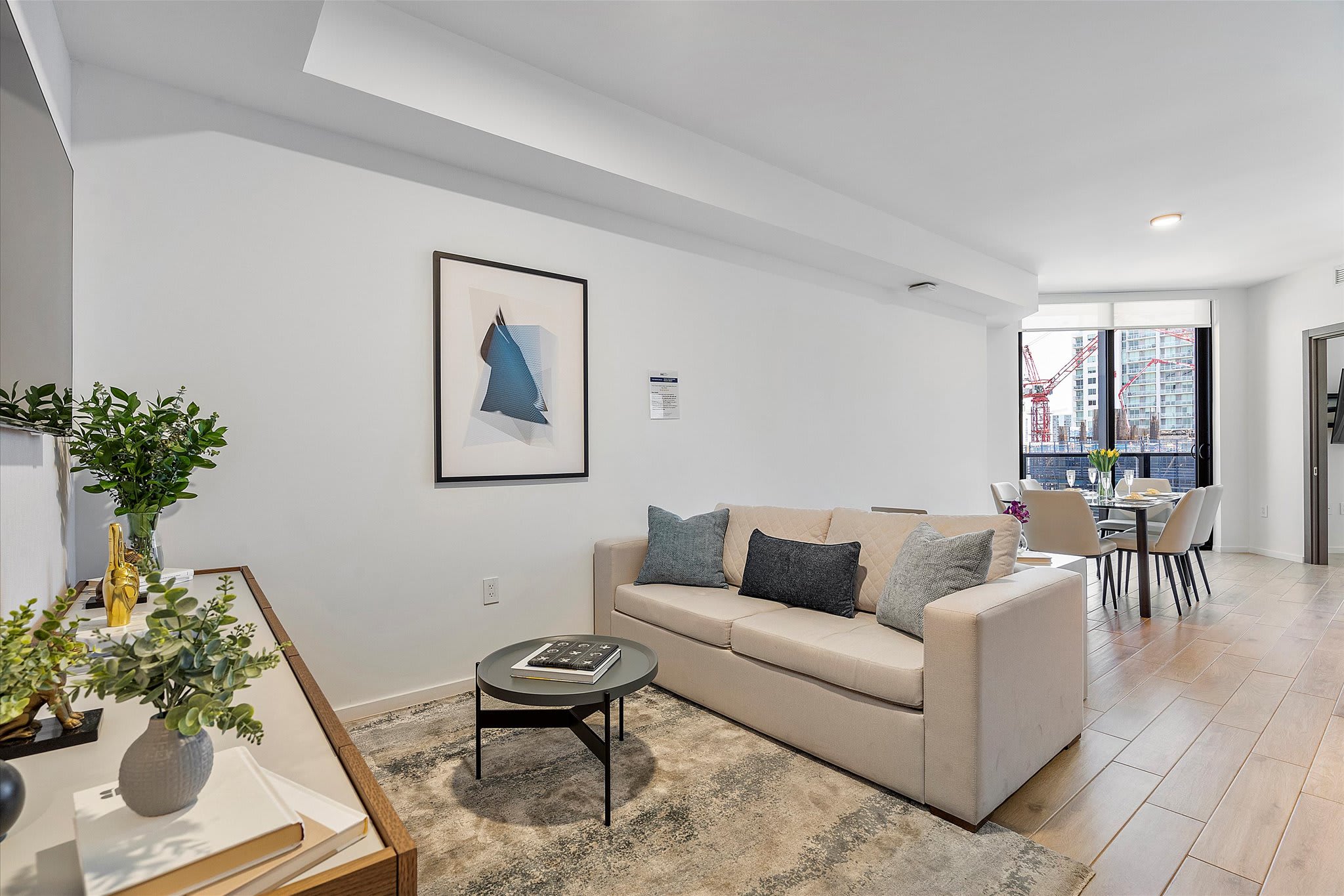 Contemporary living room with a light beige sofa, a round coffee table on a textured rug, and a large framed abstract artwork, leading into a dining area visible in the background.