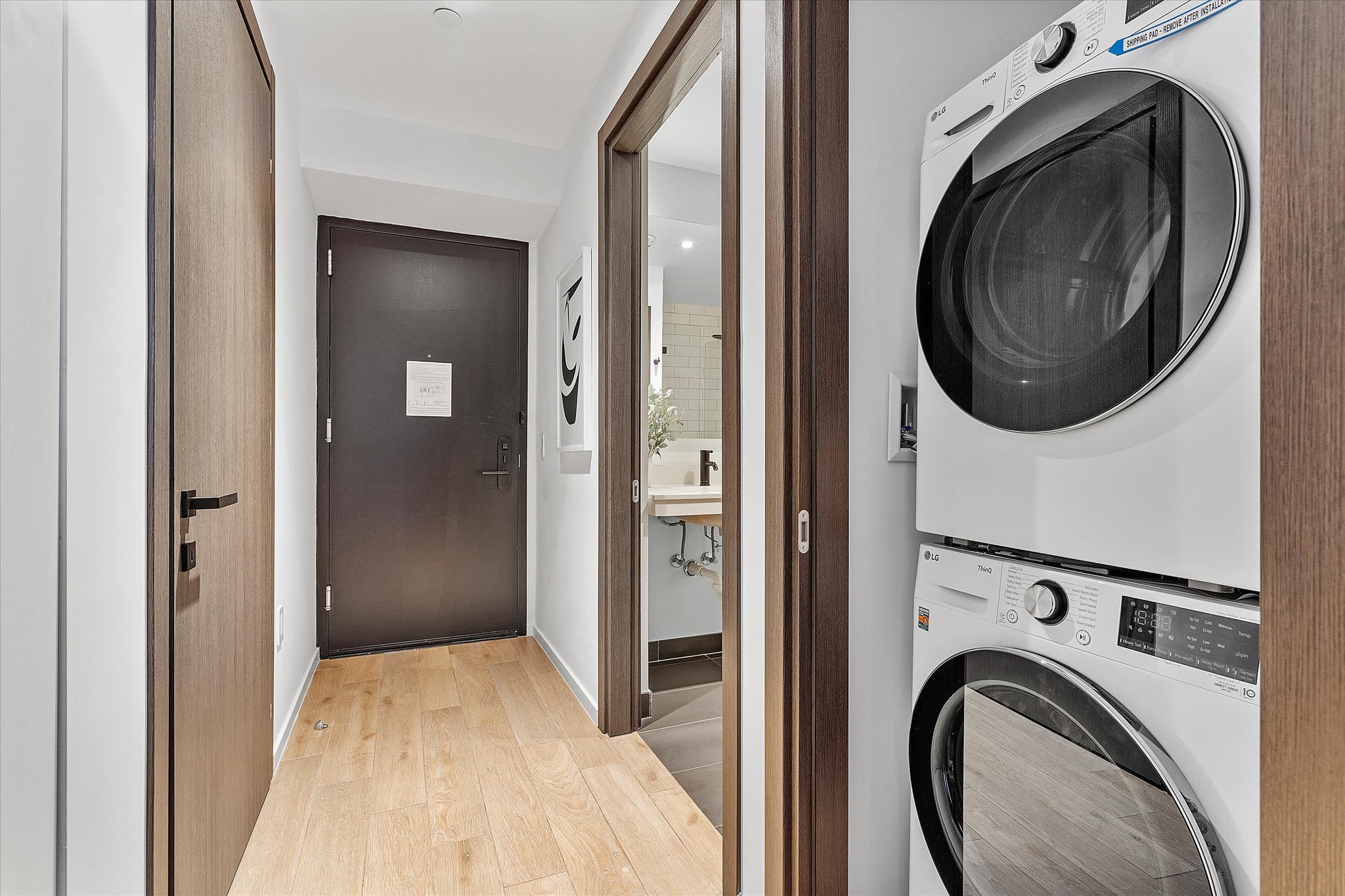 A modern interior hallway with light wood flooring, featuring a dark entry door, a stacked white washer and dryer unit, and a glimpse into a bathroom with a vanity.