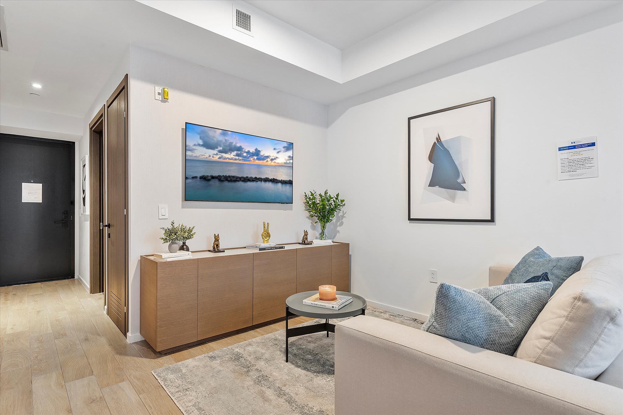 Cozy living room with a light beige sofa, a wall-mounted TV above a wooden console, and a large abstract artwork, with a view towards the entrance door.
