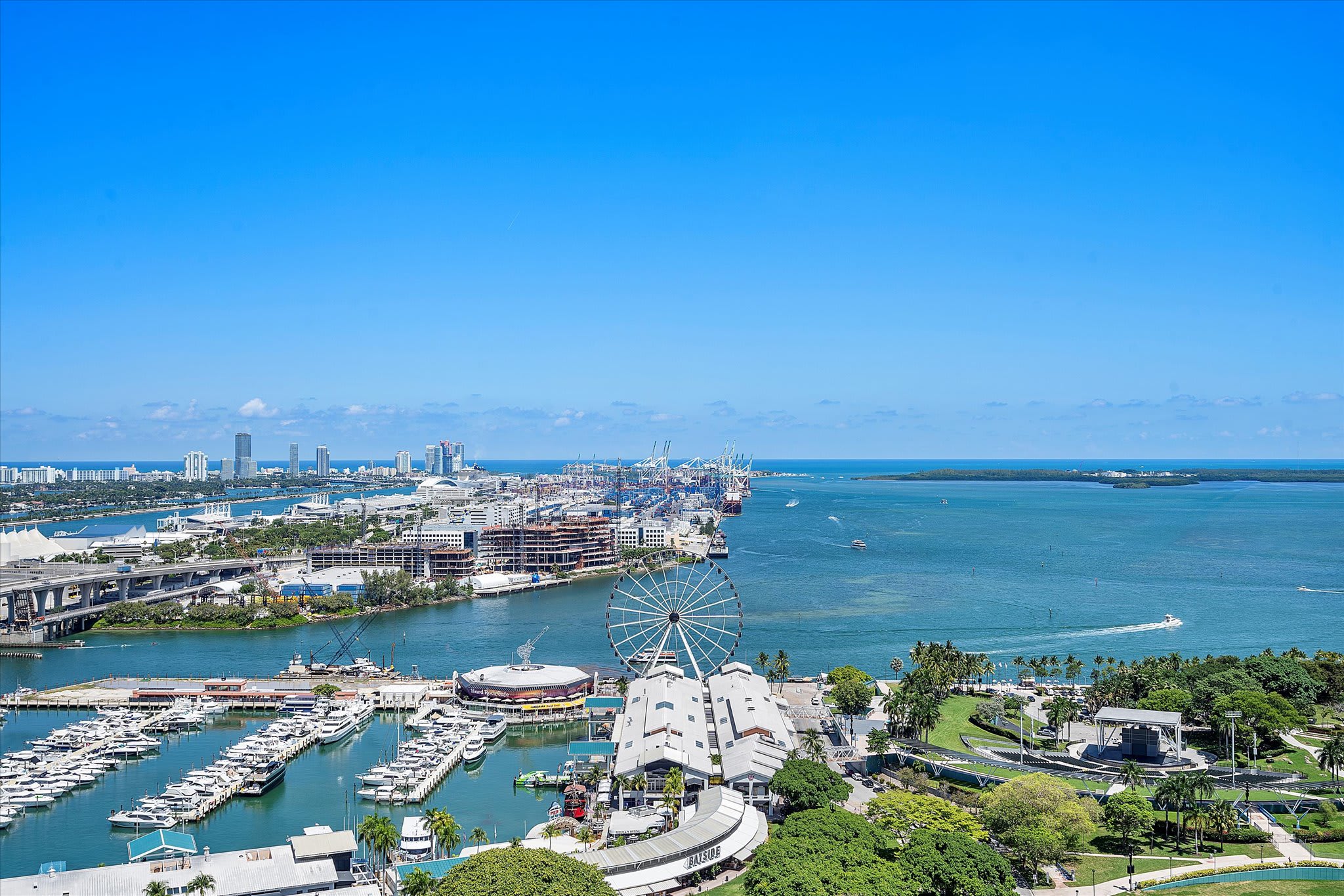 A panoramic high-angle view of a vibrant city bay, showcasing a large marina bustling with boats, a prominent Ferris wheel, and a distant urban skyline under a clear blue sky.