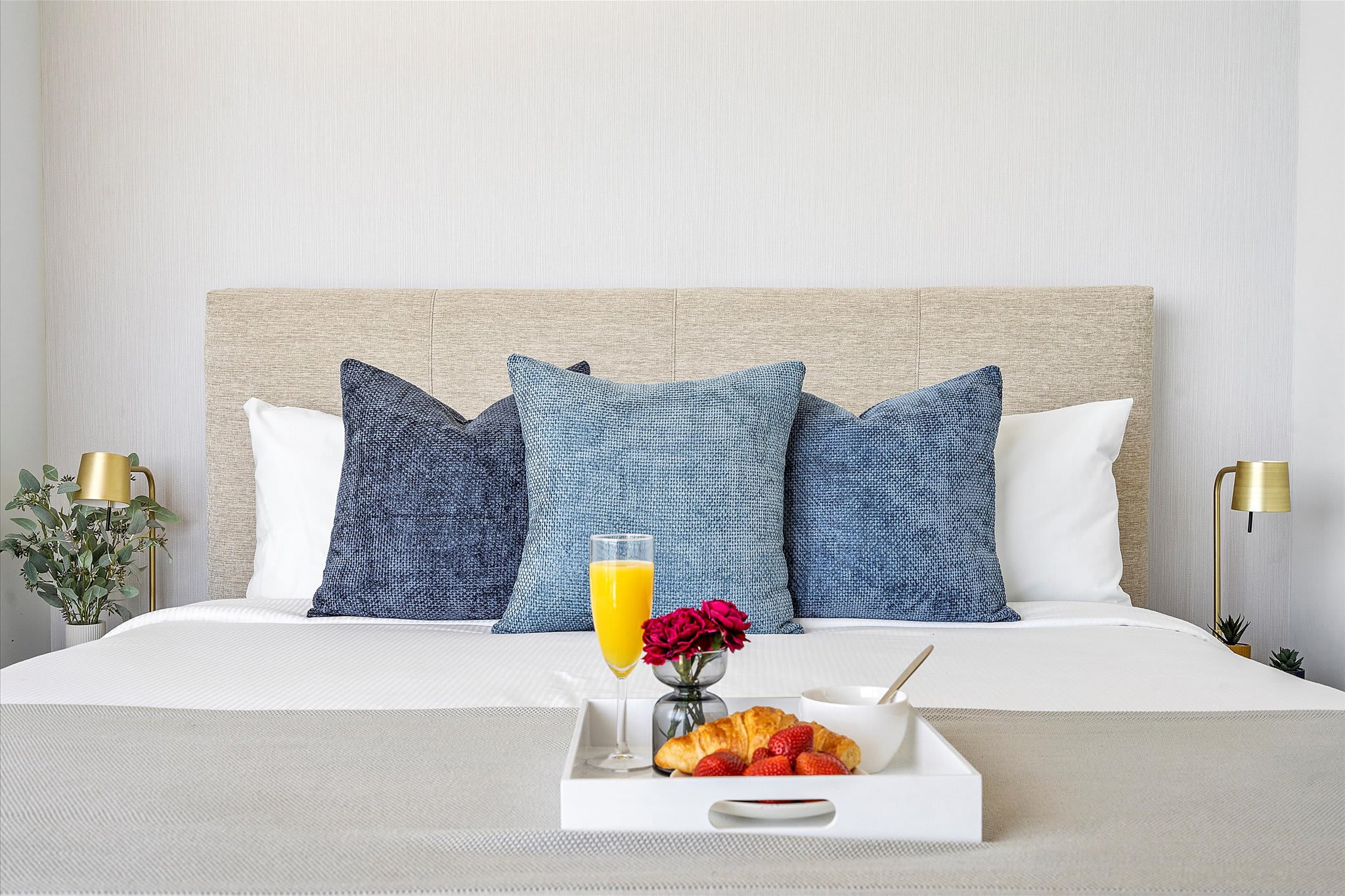 Cozy bedroom featuring a large bed with a light upholstered headboard, three blue accent pillows, and a white tray with breakfast items, flanked by modern gold-toned table lamps on nightstands.