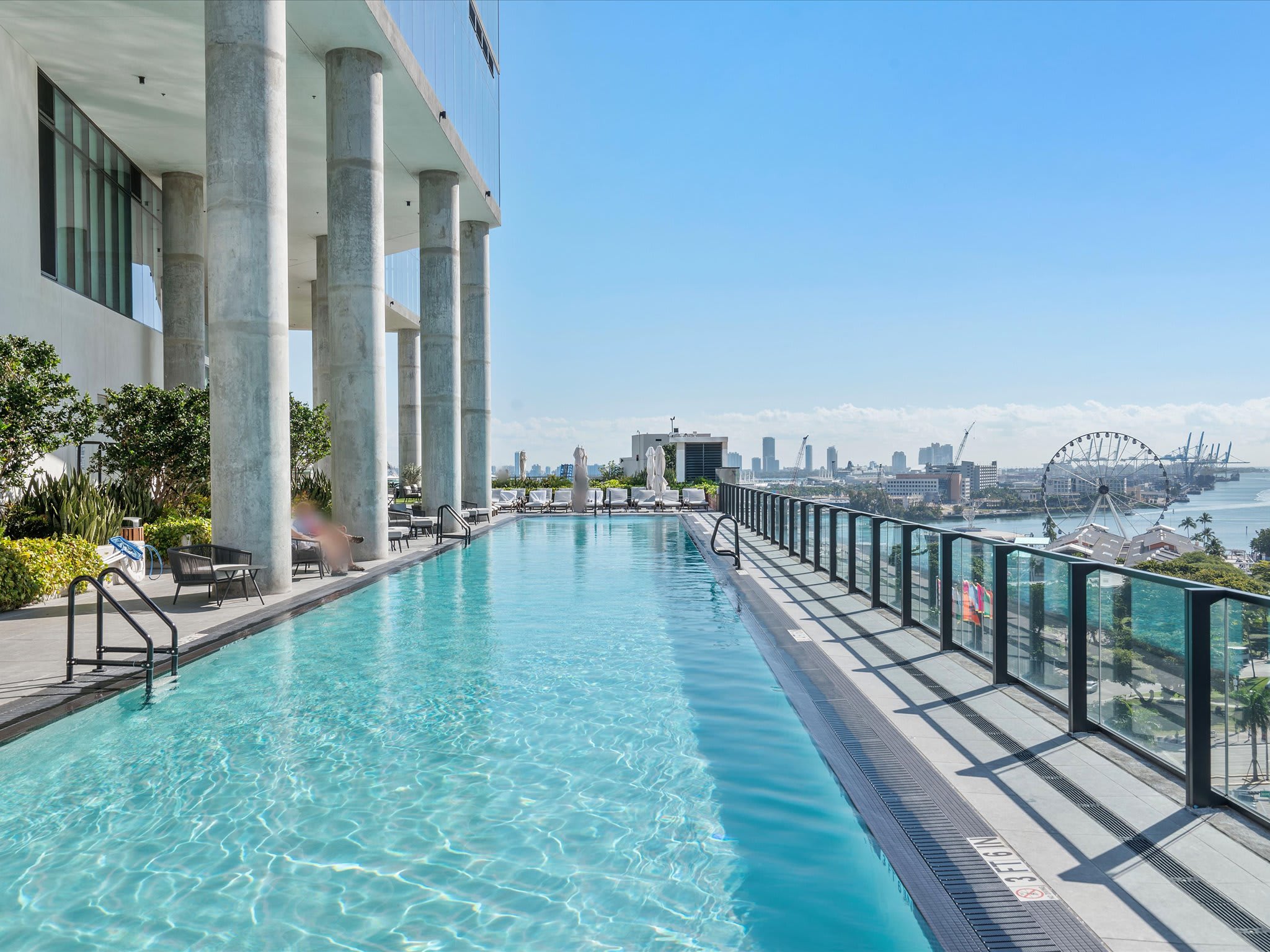 A long, narrow outdoor swimming pool on a high floor, offering expansive panoramic city and bay views. Lounge chairs and umbrellas are set up on the deck, with a glass railing providing an unobstructed vista.