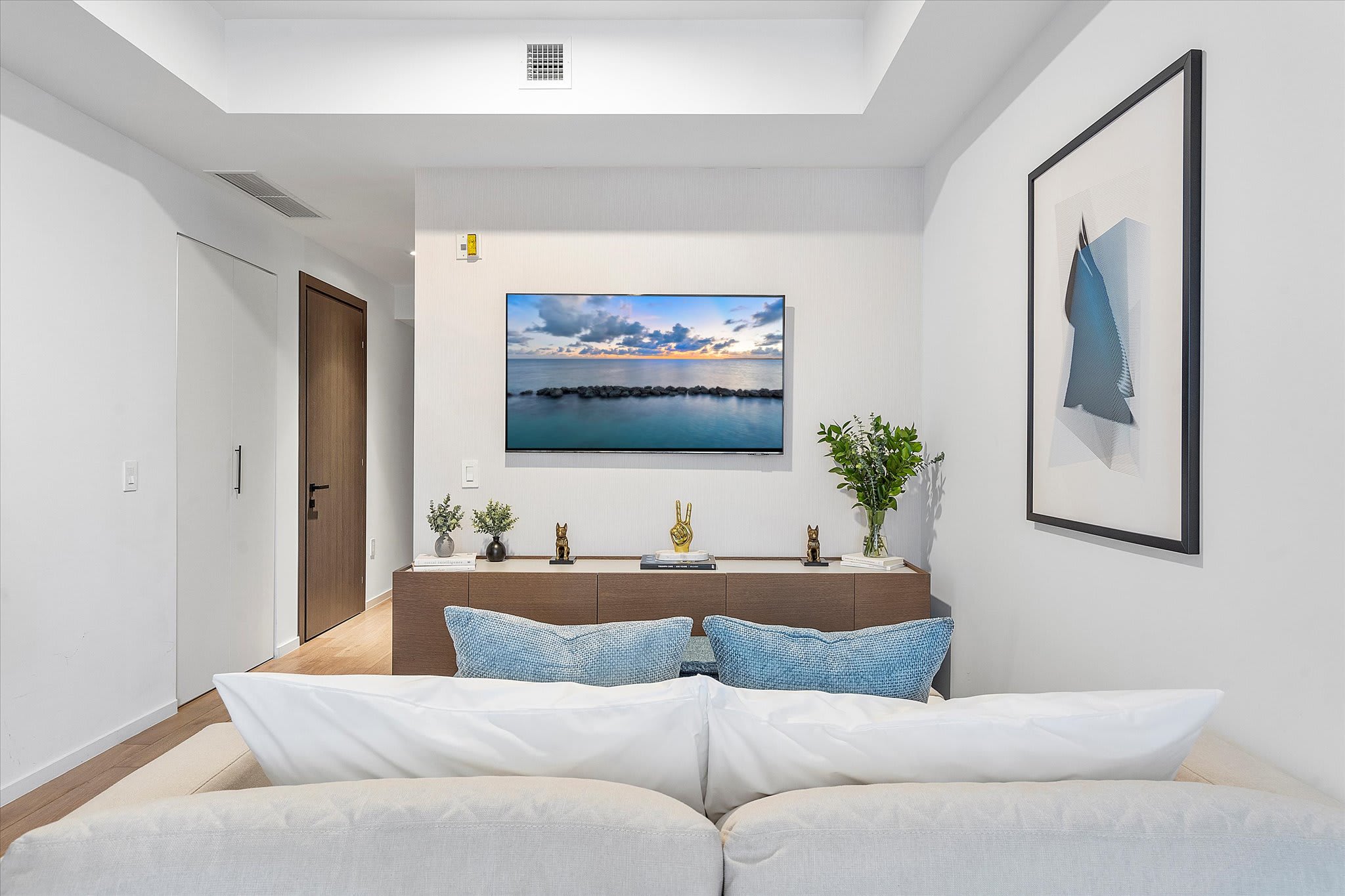 Contemporary living room featuring a light-colored sofa with blue accent pillows, a wall-mounted TV, and a wooden console table adorned with decorative items.