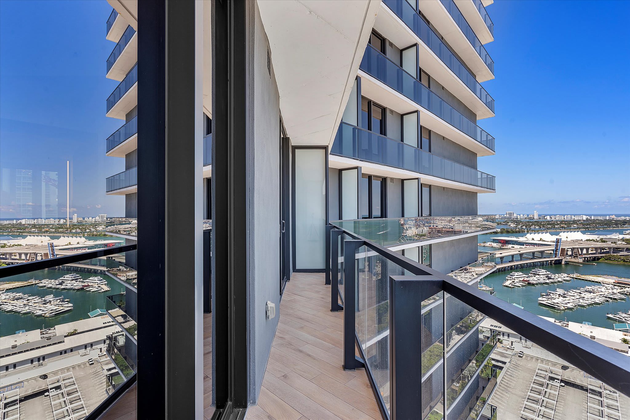 A sleek modern balcony with glass and black metal railings, light wood-look flooring, offering a stunning high-rise view of a marina filled with boats and a sprawling city skyline across the water.