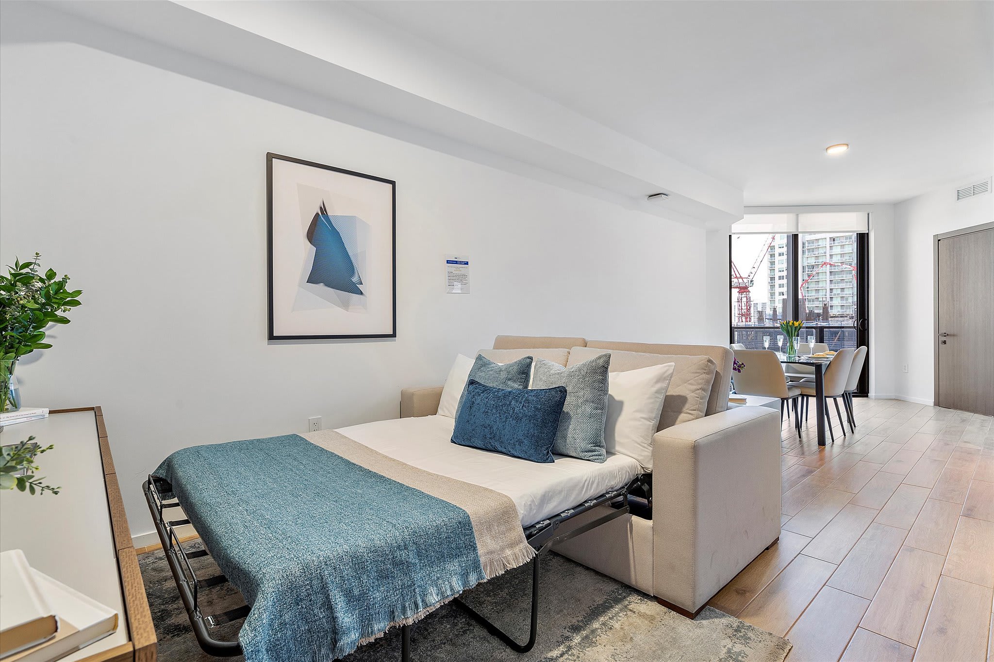 Living room transformed into a sleeping area with a pull-out sofa bed, featuring blue and white bedding, a round coffee table, and an abstract artwork on the wall.