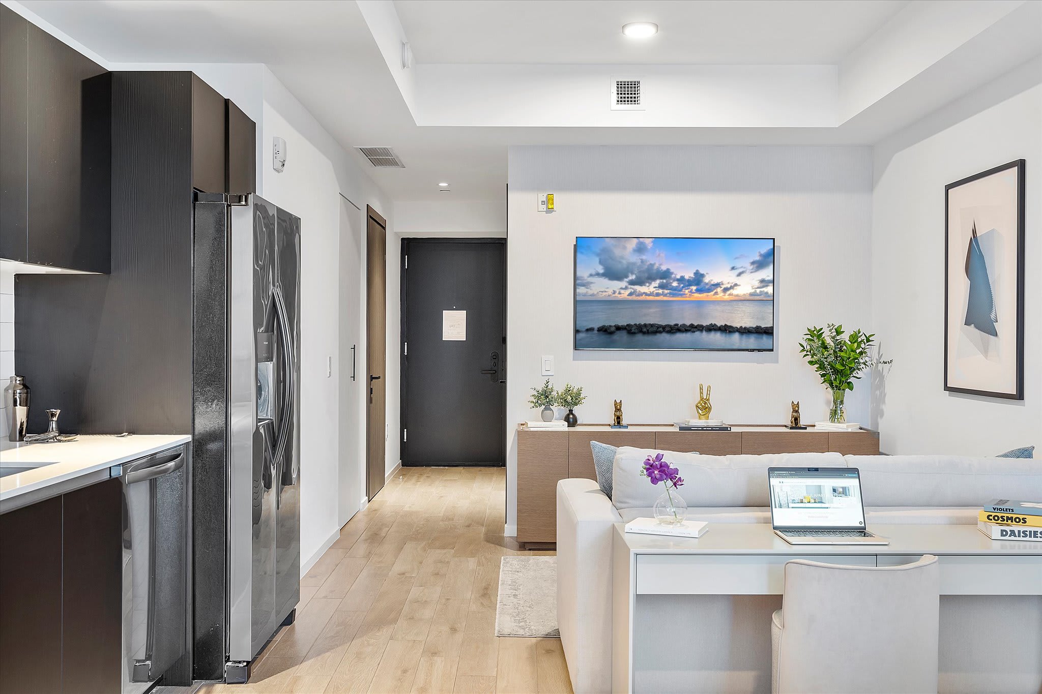Modern open-concept living area with a large wall-mounted TV displaying a seascape, a light-colored sofa, and a desk, adjacent to a dark-toned kitchen with stainless steel appliances and light hardwood floors.