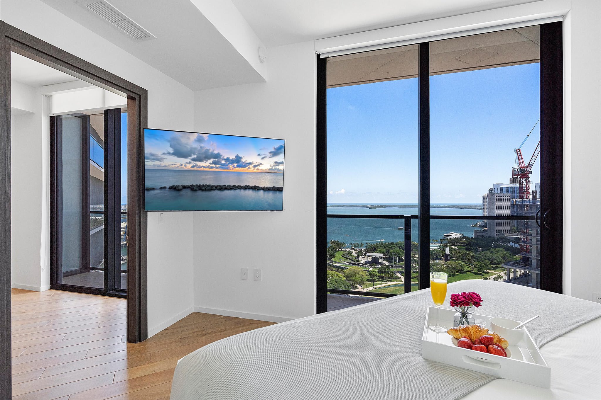 Master bedroom with a king-sized bed, a wall-mounted TV displaying a beach scene, and expansive floor-to-ceiling windows offering a panoramic city and bay view.