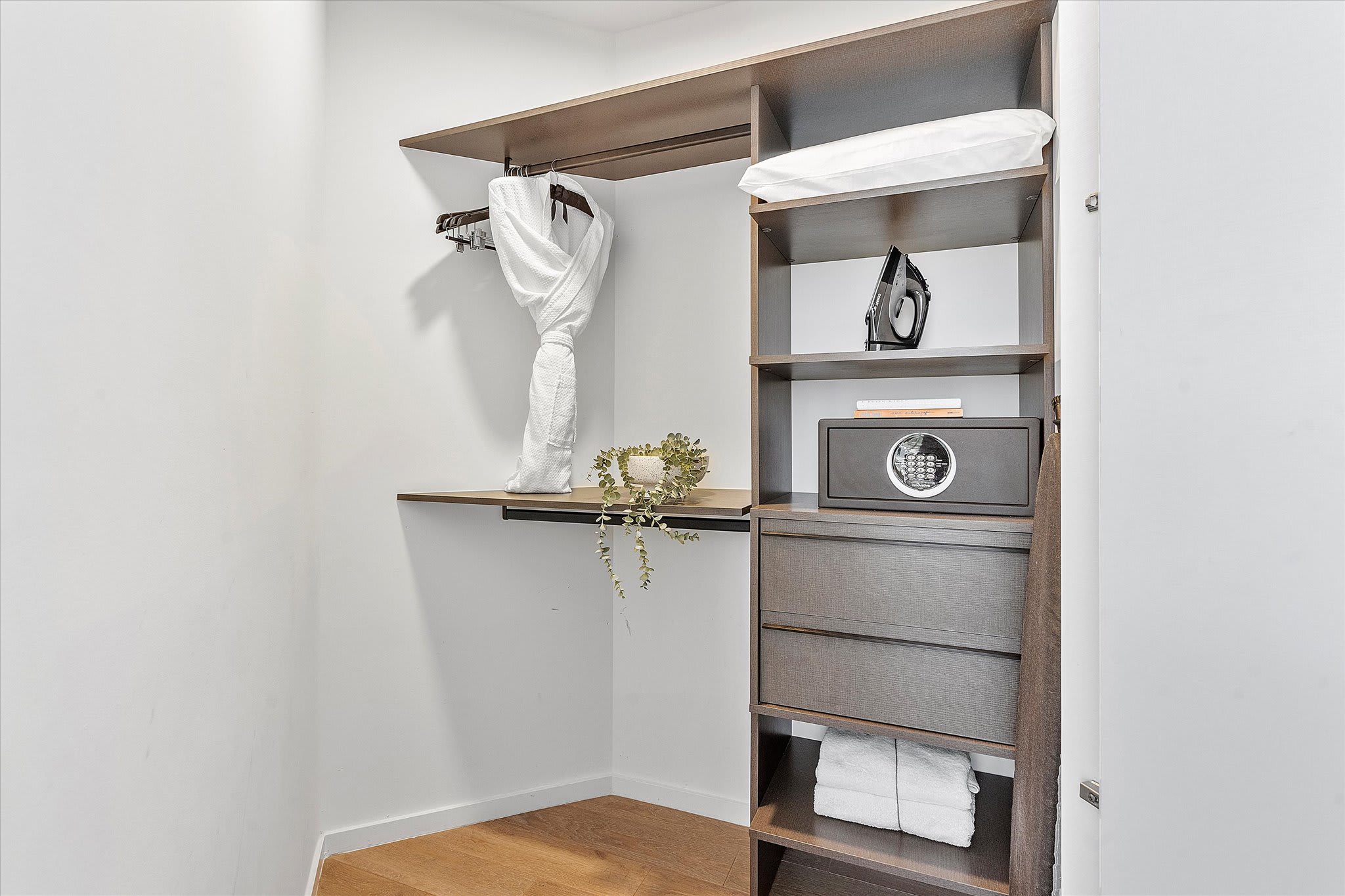 Organized walk-in closet with dark wood shelving, a hanging rod with a white robe, a safe, an iron, and neatly folded towels, providing practical storage solutions.