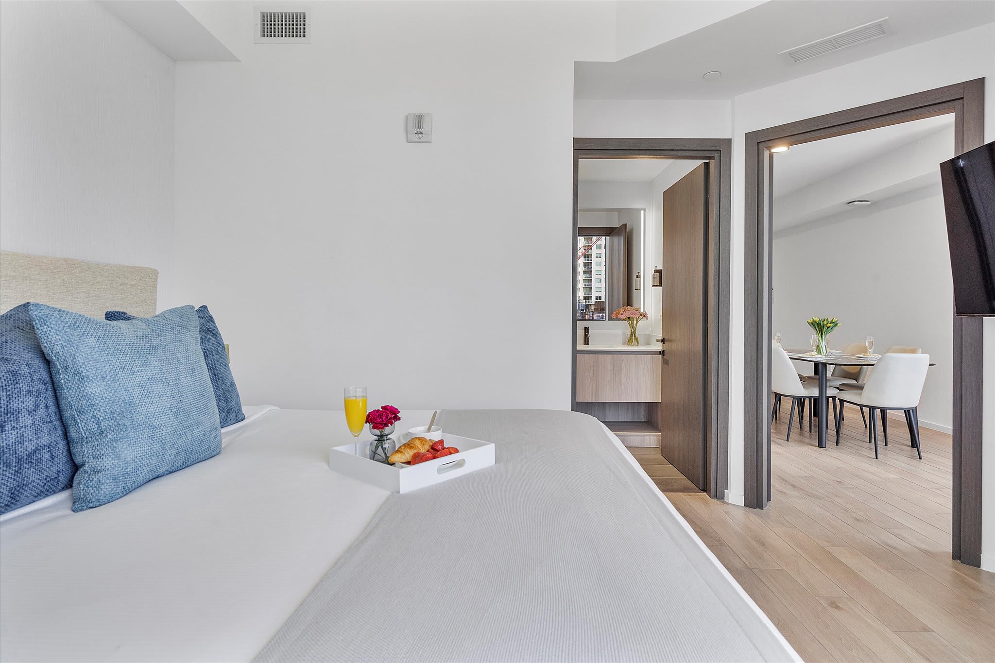 View from the bedroom showing the bed with a breakfast tray, an open doorway leading to a modern bathroom vanity, and another doorway providing access to the main living area.
