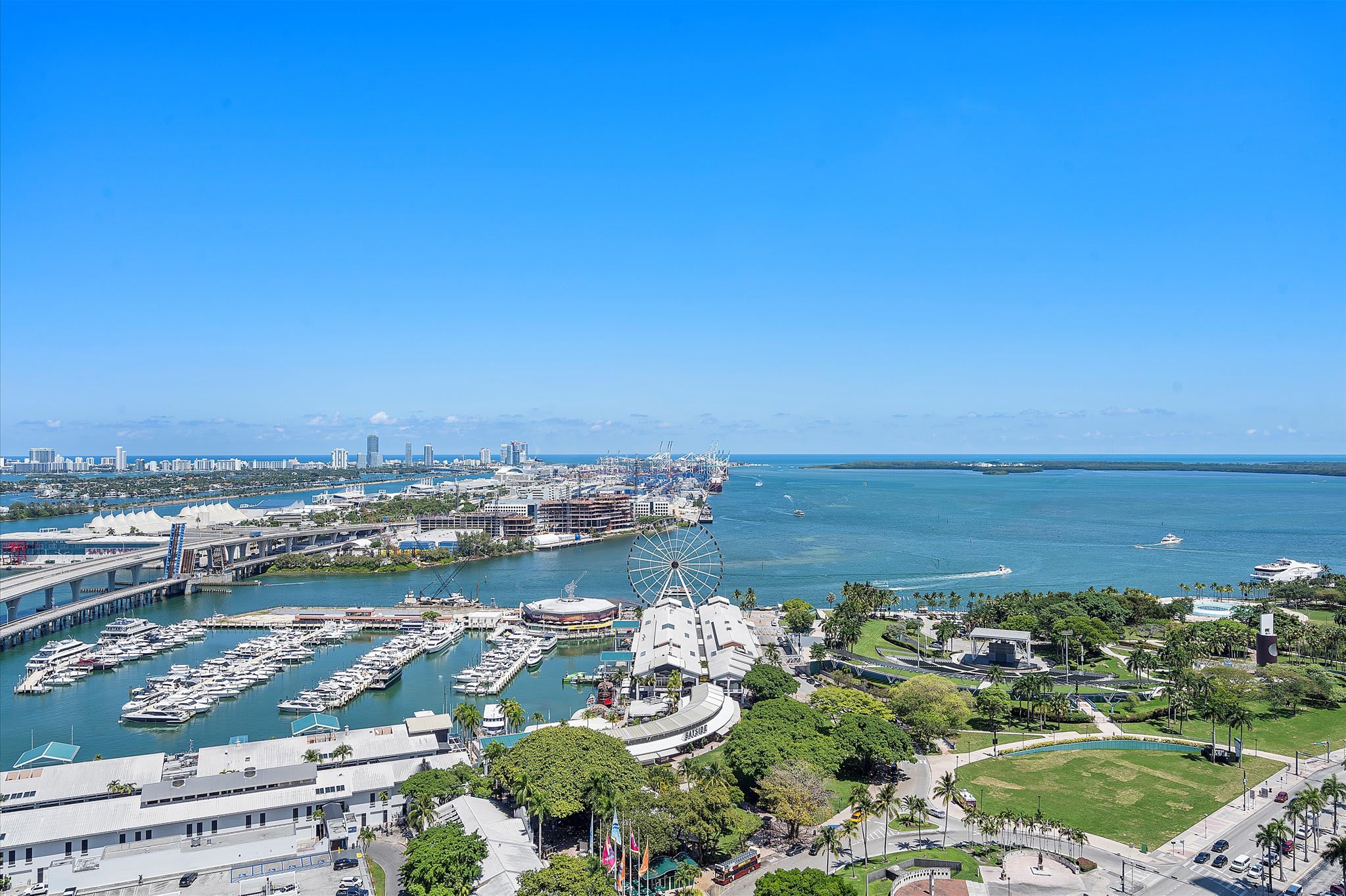 Expansive panoramic view from a high-rise balcony, showcasing a vibrant cityscape, a bustling marina filled with numerous boats, and the vast blue ocean under a clear, sunny sky.