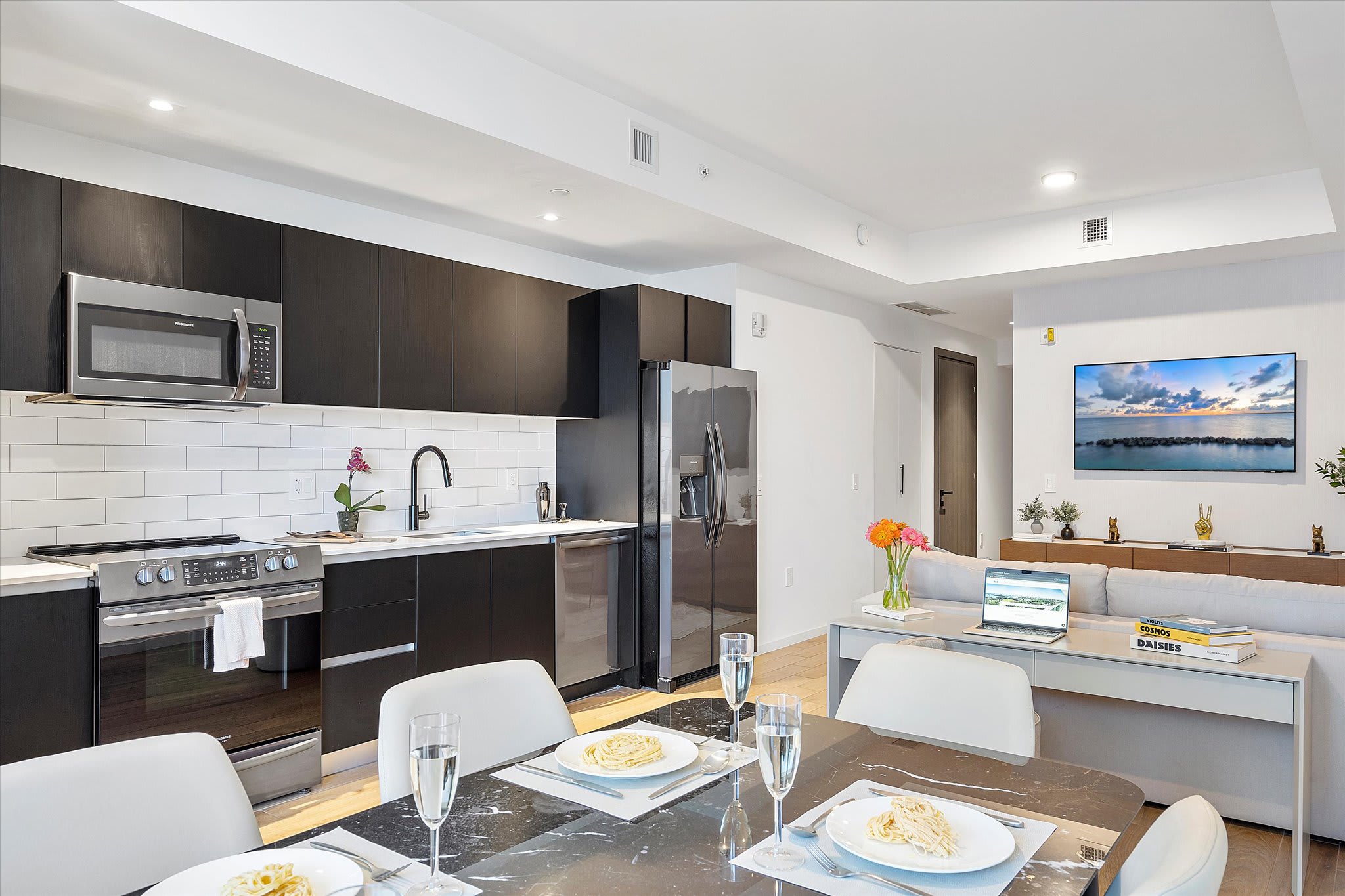 Contemporary kitchen with dark flat-panel cabinetry, white subway tile backsplash, stainless steel appliances including a gas range and microwave, and an adjacent dining table set for four with light chairs.