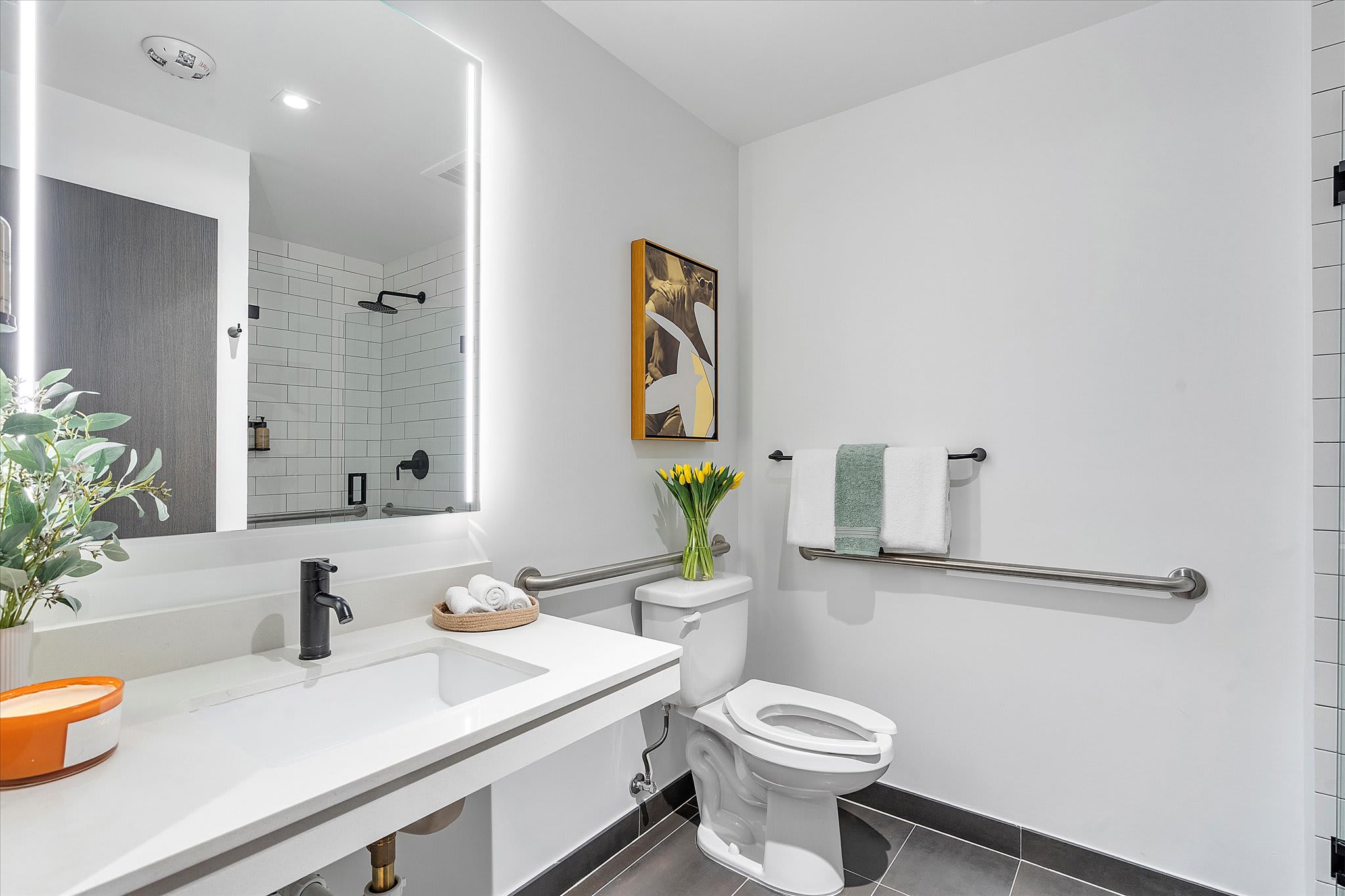 Bright modern bathroom with a white floating vanity, large illuminated mirror, a toilet with grab bars, and a subway-tiled walk-in shower visible in the mirror's reflection.