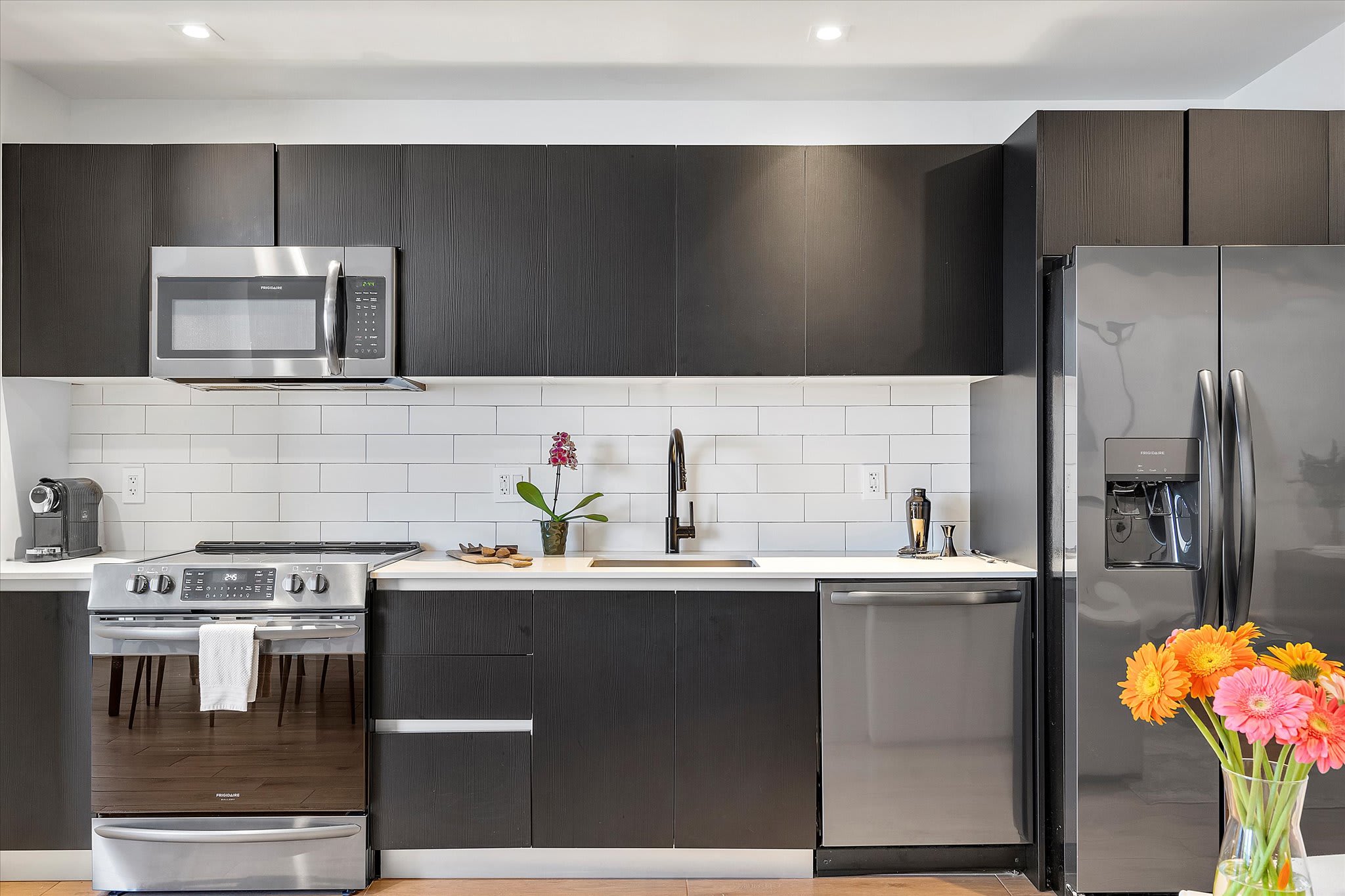 Modern kitchen with dark wood-look cabinetry, white countertops, a white subway tile backsplash, and stainless steel appliances including a microwave, oven, and double-door refrigerator.