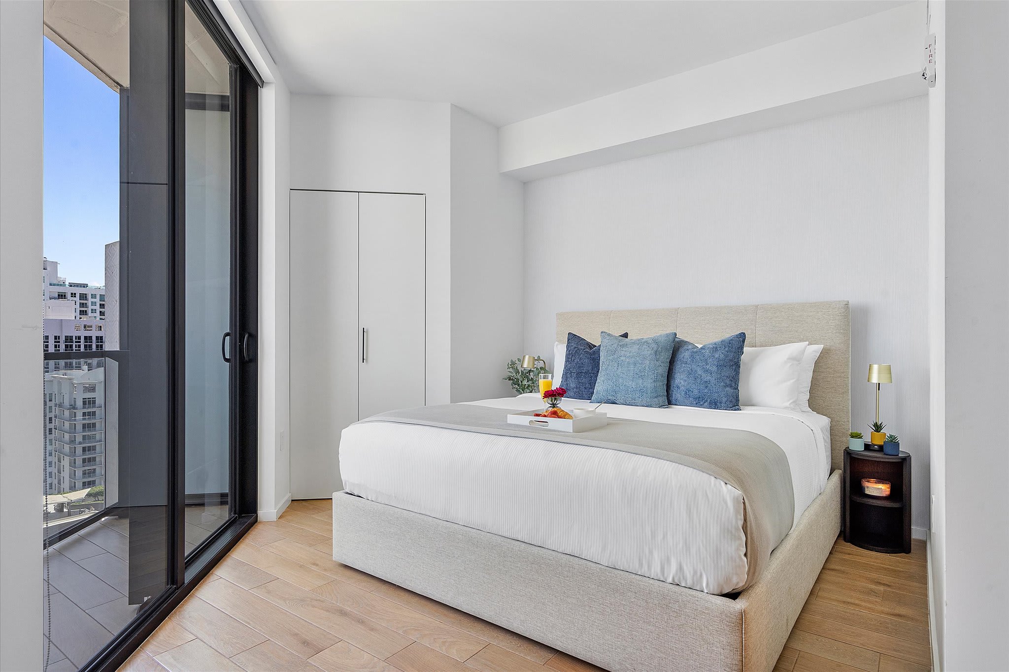 Bright bedroom with a king-sized bed featuring a light upholstered headboard and blue accent pillows, a small nightstand, and a large sliding glass door leading to a balcony.