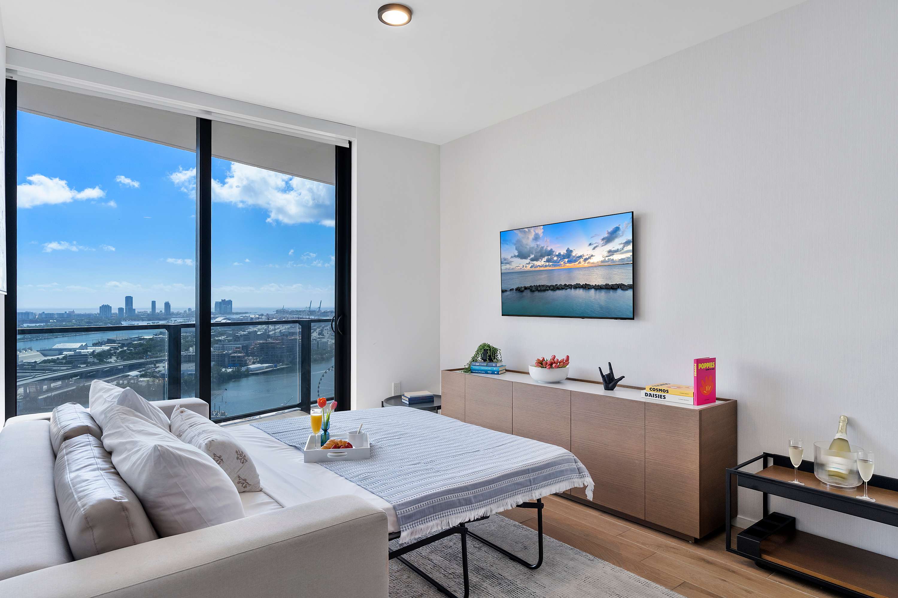 A contemporary living space with a pull-out sofa bed, a long wooden console table, and a wall-mounted TV, all illuminated by natural light from expansive windows showcasing a city and water panorama.