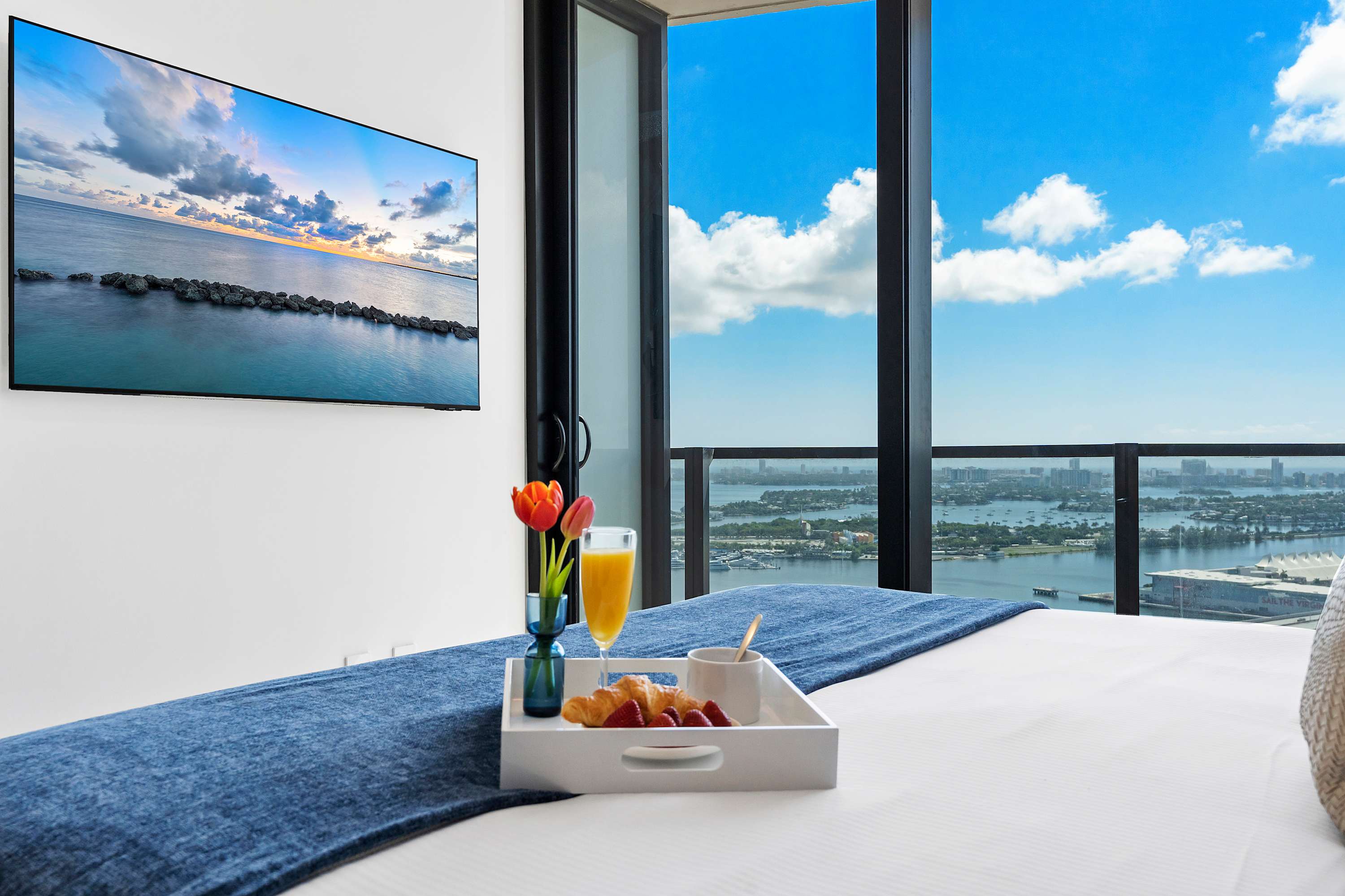 A bright bedroom featuring a white bed with a blue throw, a breakfast tray, and a wall-mounted TV, all set against a large window offering panoramic city and water views under a blue sky.