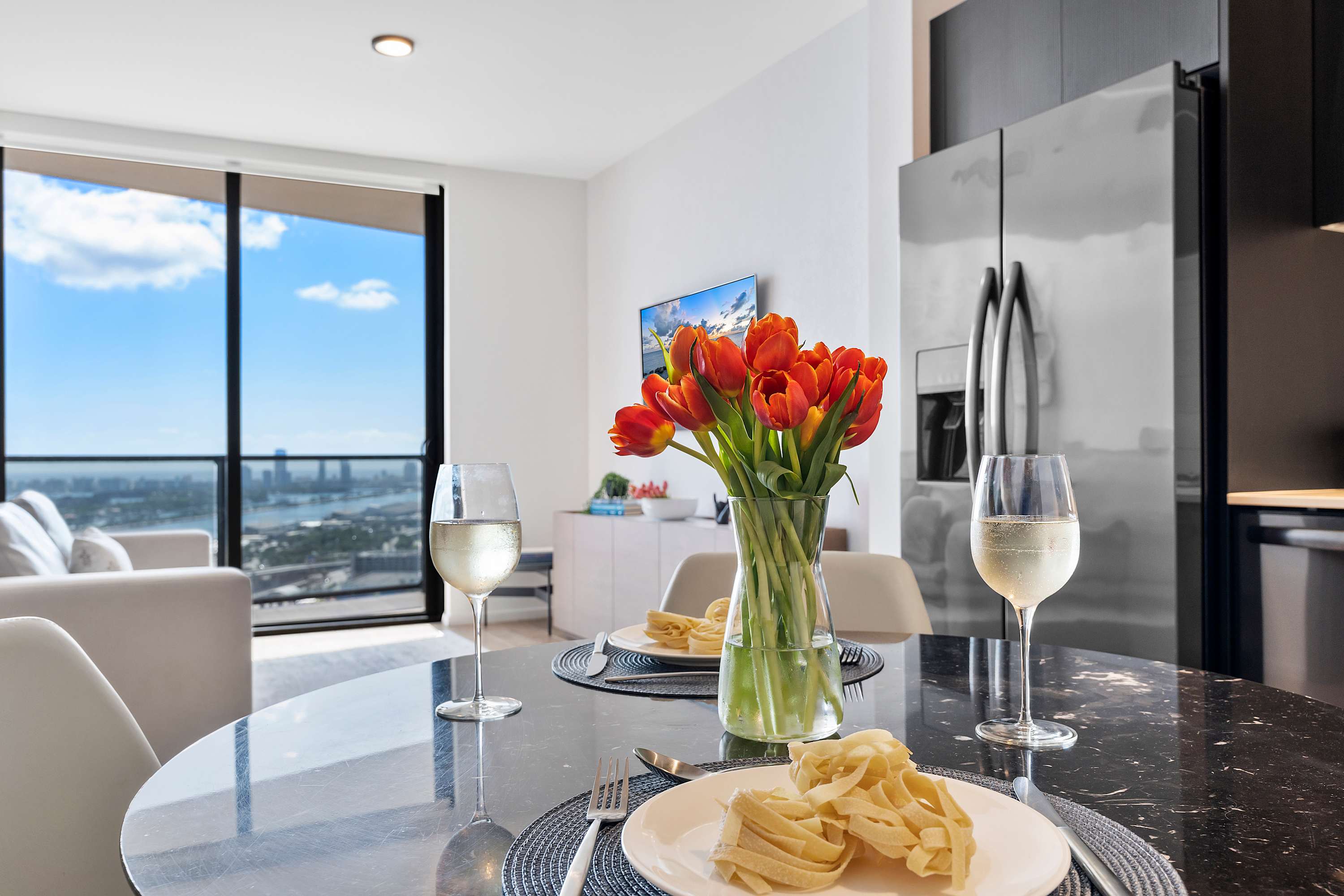 Modern dining area features a round black marble table set with two places, white wine, and a vase of orange tulips, with a sleek kitchen and city views through large windows in the background.
