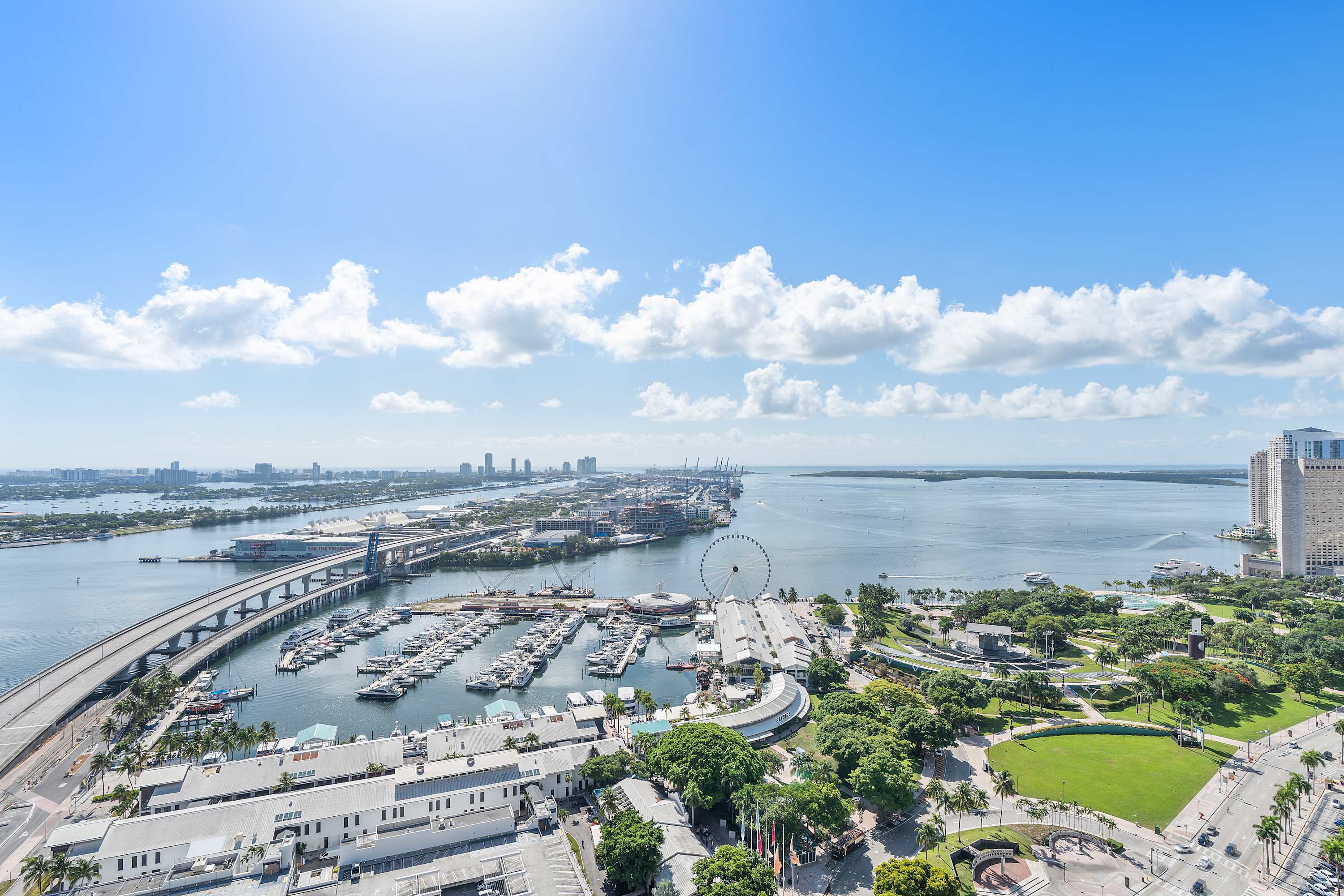 High-angle aerial view captures a bustling marina filled with numerous boats, adjacent to a large Ferris wheel, surrounded by urban infrastructure, bridges, and green park areas under a clear sky.